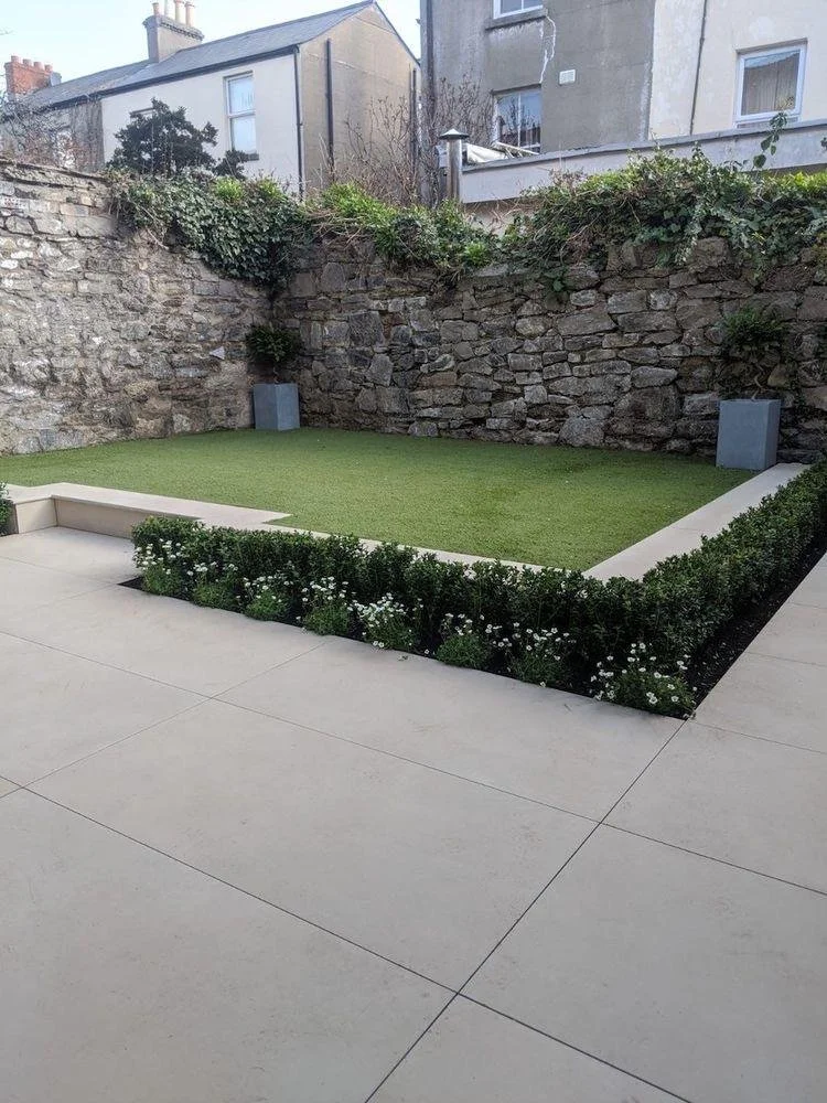 Small backyard with a patch of artificial grass, surrounded by a low white border and flower bed with small white flowers. The yard is enclosed by a stone wall and neighboring buildings are visible in the background.