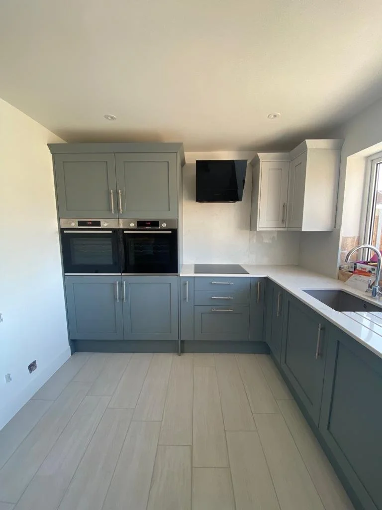 Modern kitchen with gray cabinets, built-in ovens, black range hood, and a large window with a white countertop and sink.
