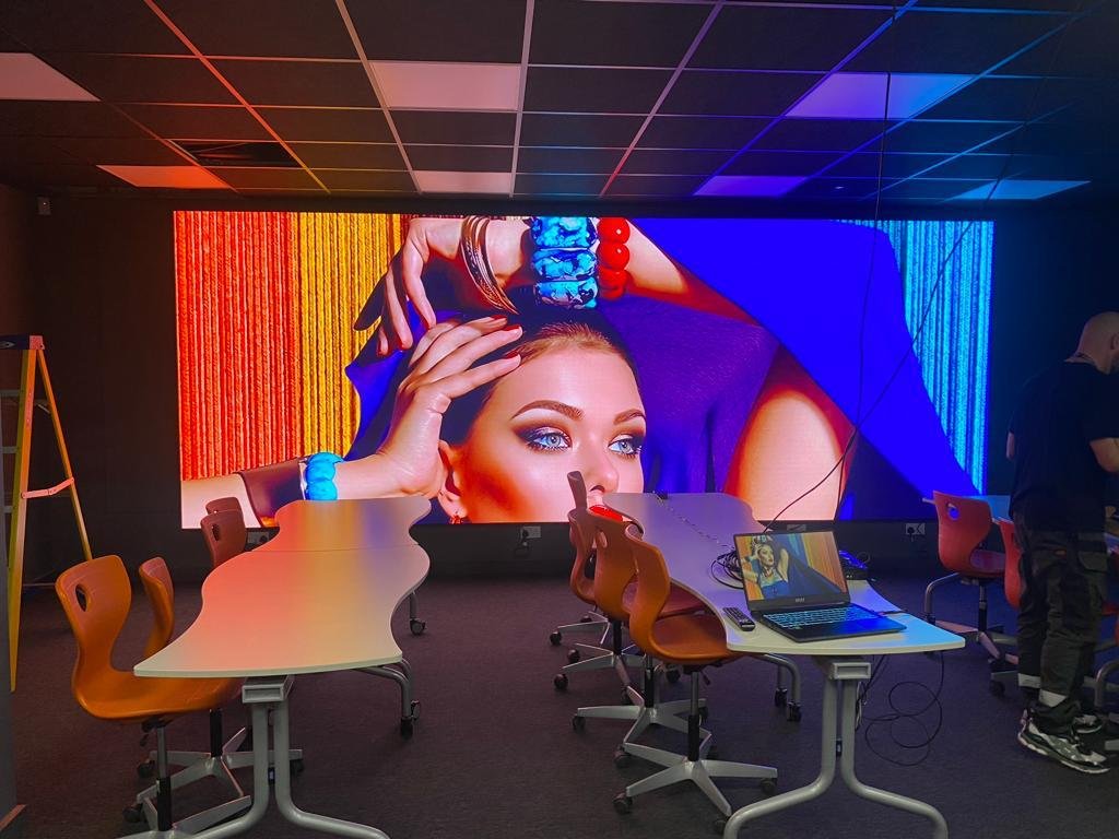 A large digital screen displays a close-up of a woman's face with makeup, jewelry, and striking eye makeup, in a modern room with tables, chairs, and a laptop.