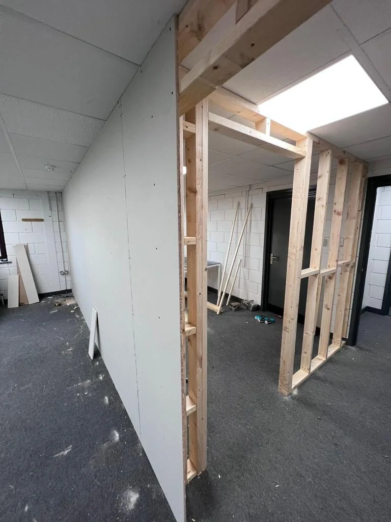 Interior of a room under construction with a partially built wall made of wooden studs and drywall. The room has white brick walls, ceiling tiles, and a dark door, with construction tools and materials scattered around.
