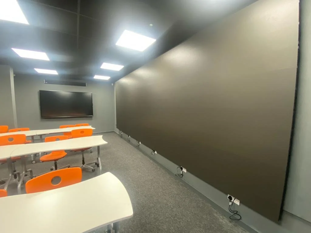 Empty classroom with white tables and orange chairs, large black wall-mounted screen, dark gray wall, gray carpet, and ceiling lights.