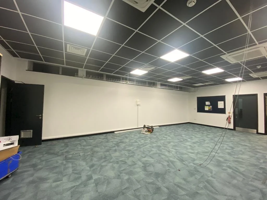 Empty room with a black-and-white checkered ceiling, fluorescent lights, and a blue patterned carpet. There are black doors on the walls, some wiring hanging from the ceiling, and some equipment on the floor.