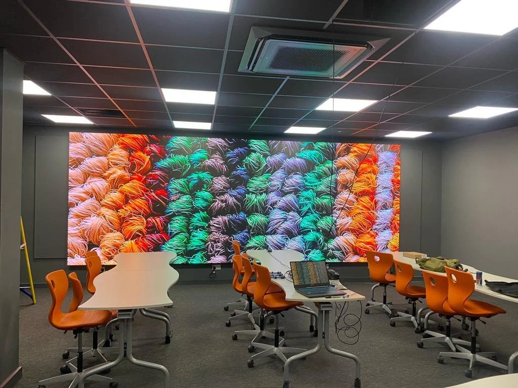 Conference room with a large colorful LED display showing yarn balls in multiple colors, orange chairs, and a table with a laptop and other items.