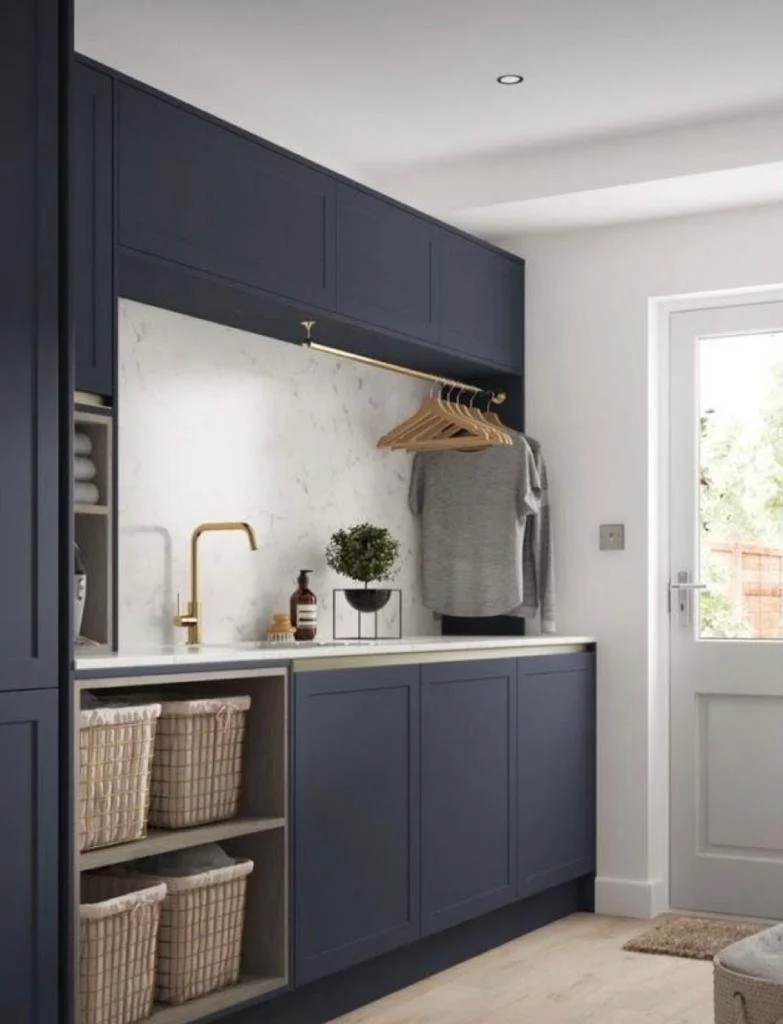 A laundry area with dark blue cabinets, a white marble countertop, a gold faucet, a small potted plant, hanging wooden hangers, a gray t-shirt, and a door leading outside.
