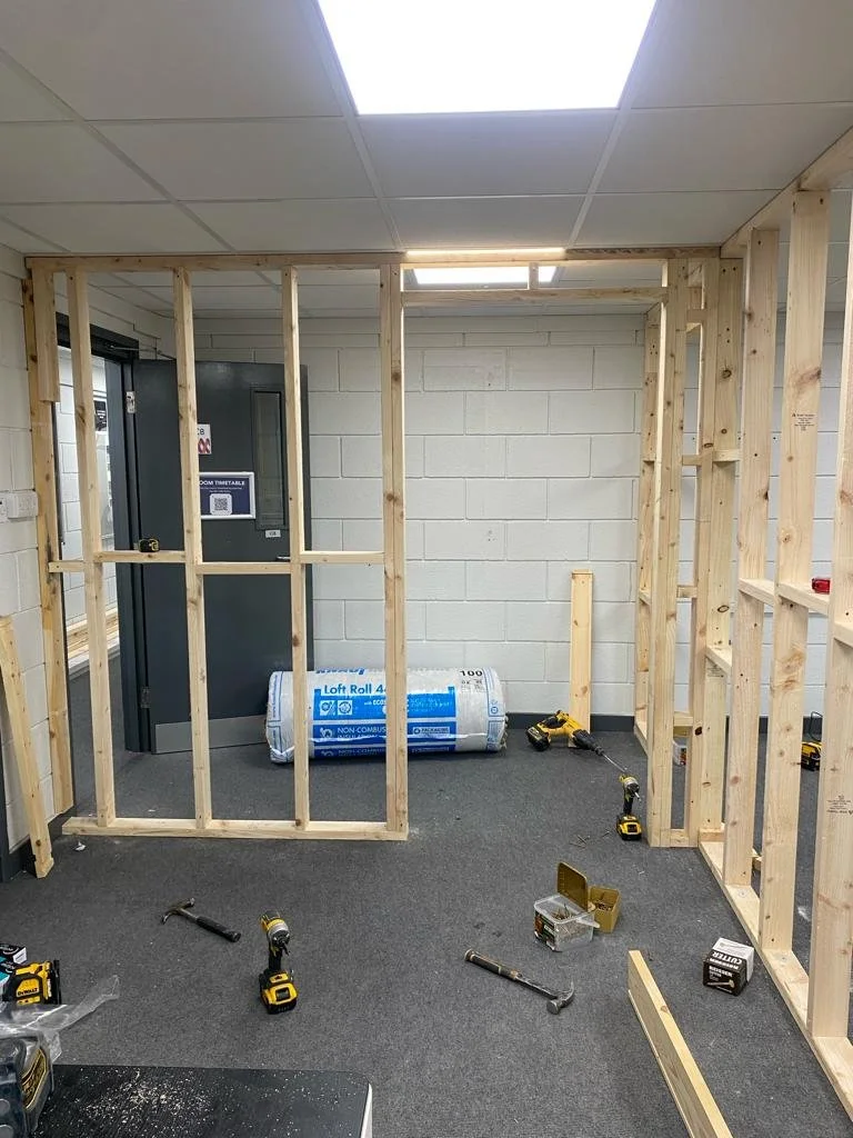 Interior of a building under construction with wooden framing, tools, a rolled insulation, and a gray door in the background.