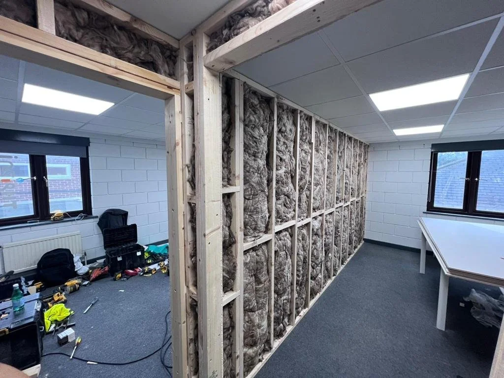 Interior of a room under construction with exposed insulation on the wall, wooden framing, and construction tools on the floor.