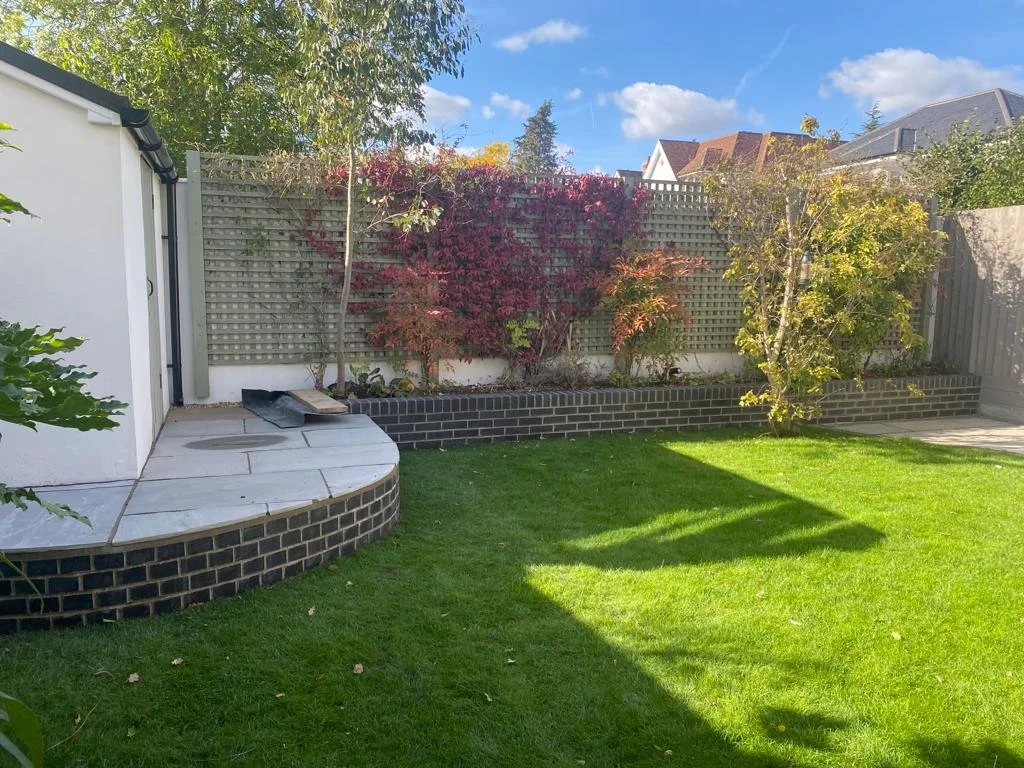 Well-maintained backyard with green grass, a raised garden bed with plants, and a wooden fence with climbing plants and trees under a blue sky.