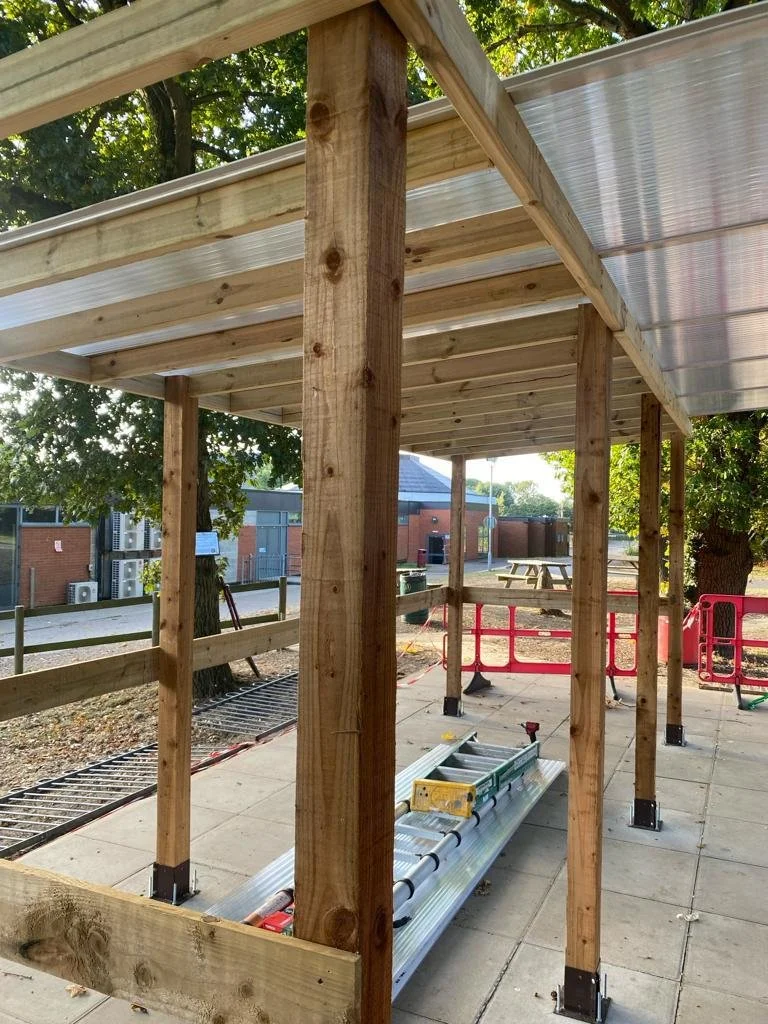 A construction site with wooden support beams and a metal roof, set on a paved surface, with tools and construction materials around.