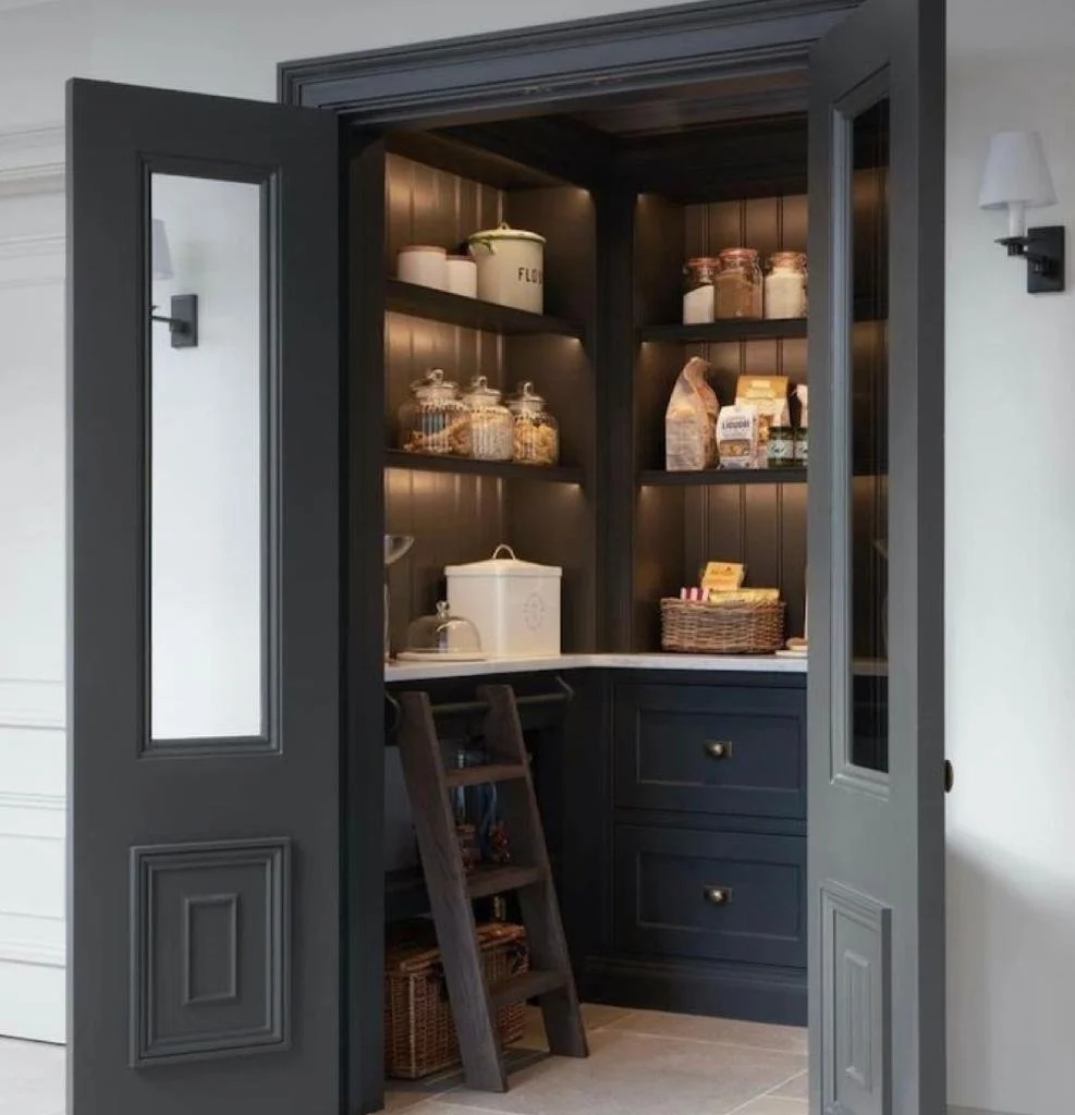 A pantry closet with dark wooden cabinets and open double doors, containing jars, boxes, cookies, and pantry staples, with a small counter space and a ladder.