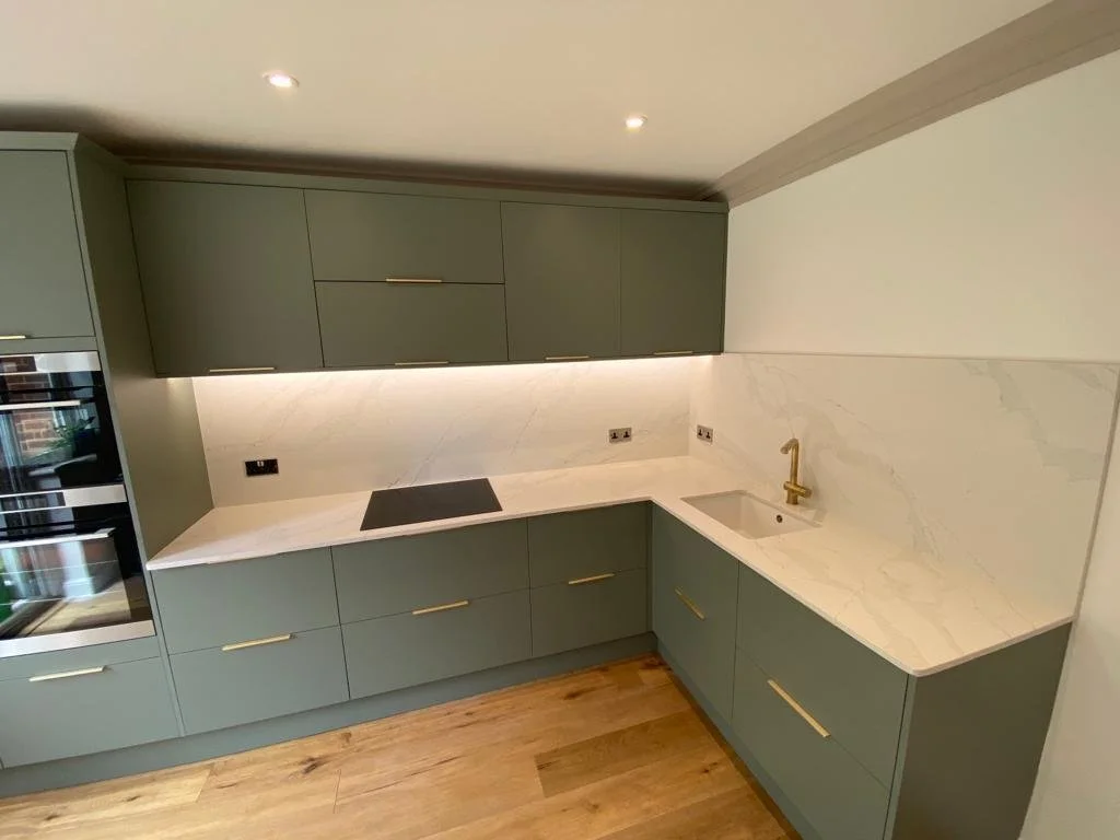 Modern kitchen with green cabinets, marble countertop and backsplash, built-in oven, black cooktop, brass handles, and a brass faucet