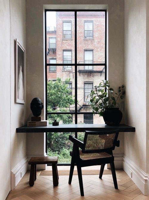 A small workspace by a large window revealing a cityscape with brick buildings and trees, featuring a black desk, a wooden stool, a potted plant, and a black sculpture.