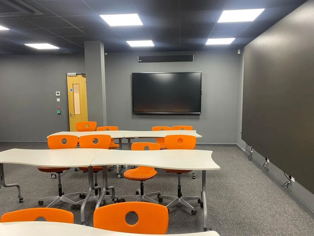 Empty classroom with white tables, orange chairs, a large black screen, gray walls, and ceiling lights.
