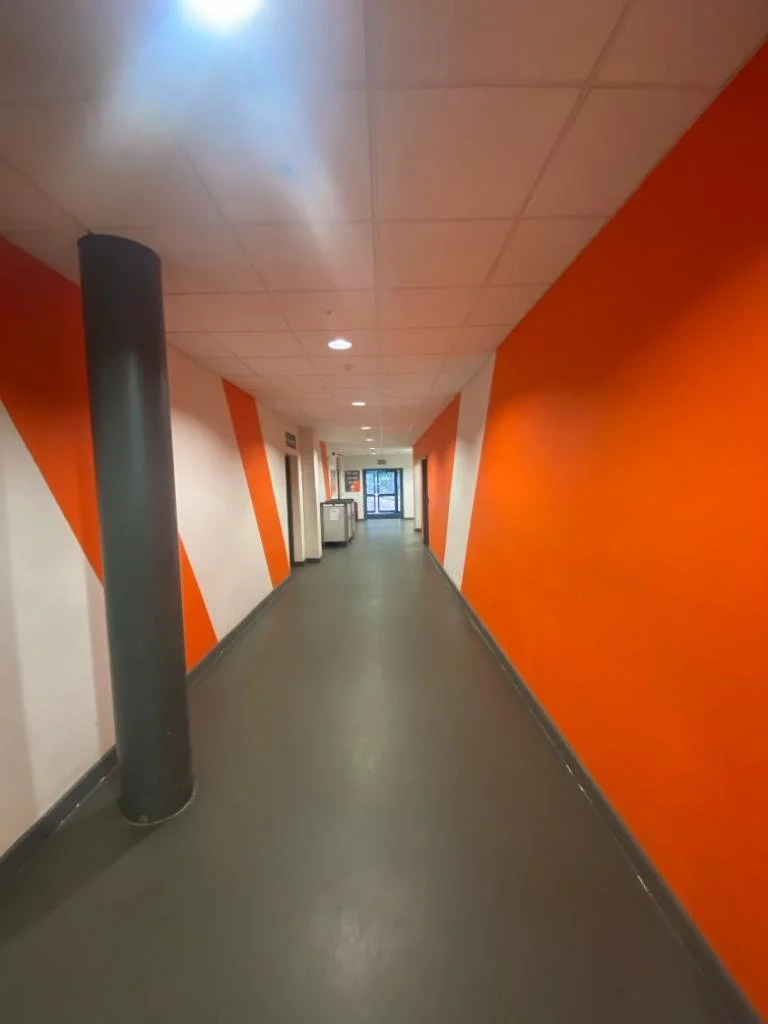 Empty hallway with orange and white walls, gray flooring, ceiling lights, and a glass door at the end.