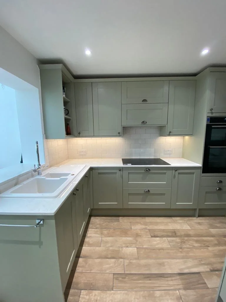 Kitchen with light green cabinets, white subway tile backsplash, white countertop, double sink, electric cooktop, built-in oven, and wood-look flooring.