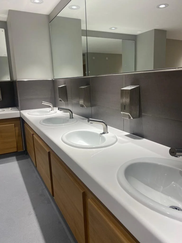 Public restroom with three white sinks and mirrors above, stainless steel soap dispensers on the wall, wooden cabinets below, and a gray tiled wall.