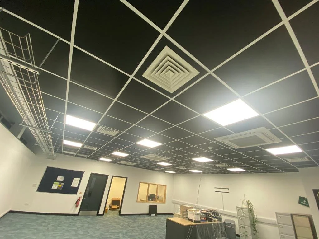 Empty office space with black suspended ceiling and fluorescent lights, some with vents, a black door, and unfinished furniture.