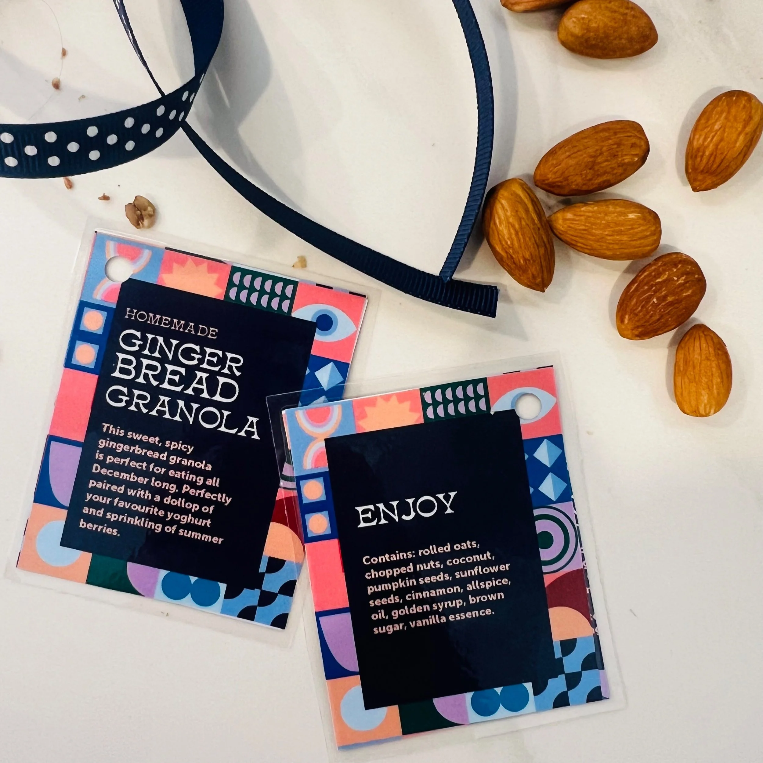 Two colorful tags for homemade gingerbread granola, one labeled "Ginger Bread Granola" and the other labeled "Enjoy," with almonds, a navy polka dot ribbon, and some loose oats and nuts around them.