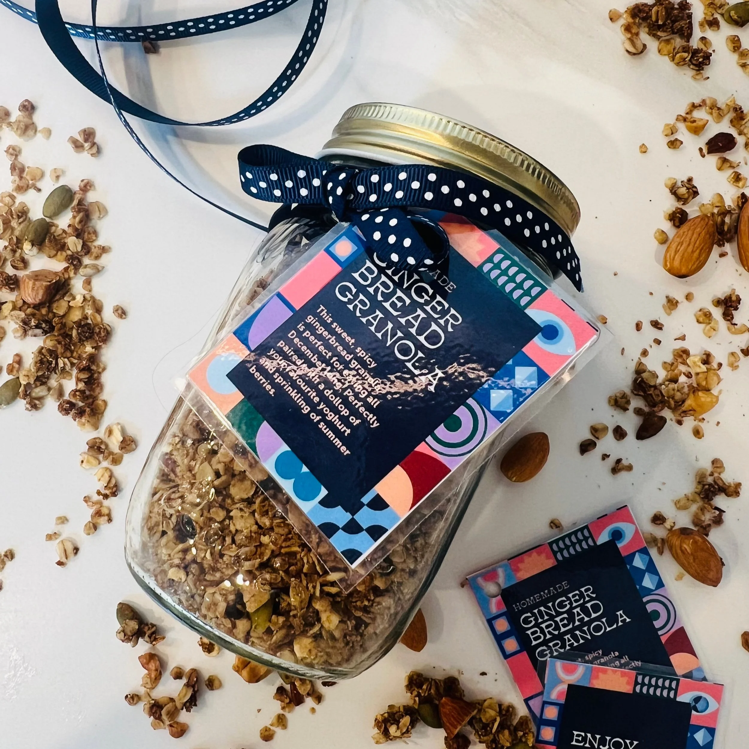 A glass jar labeled gingerbread granola with a dark blue and colorful geometric patterned label, tied with a blue polka dot ribbon, resting on a white surface. Around the jar are scattered granola clusters and whole almonds, with a packaged gingerbread granola treat nearby.