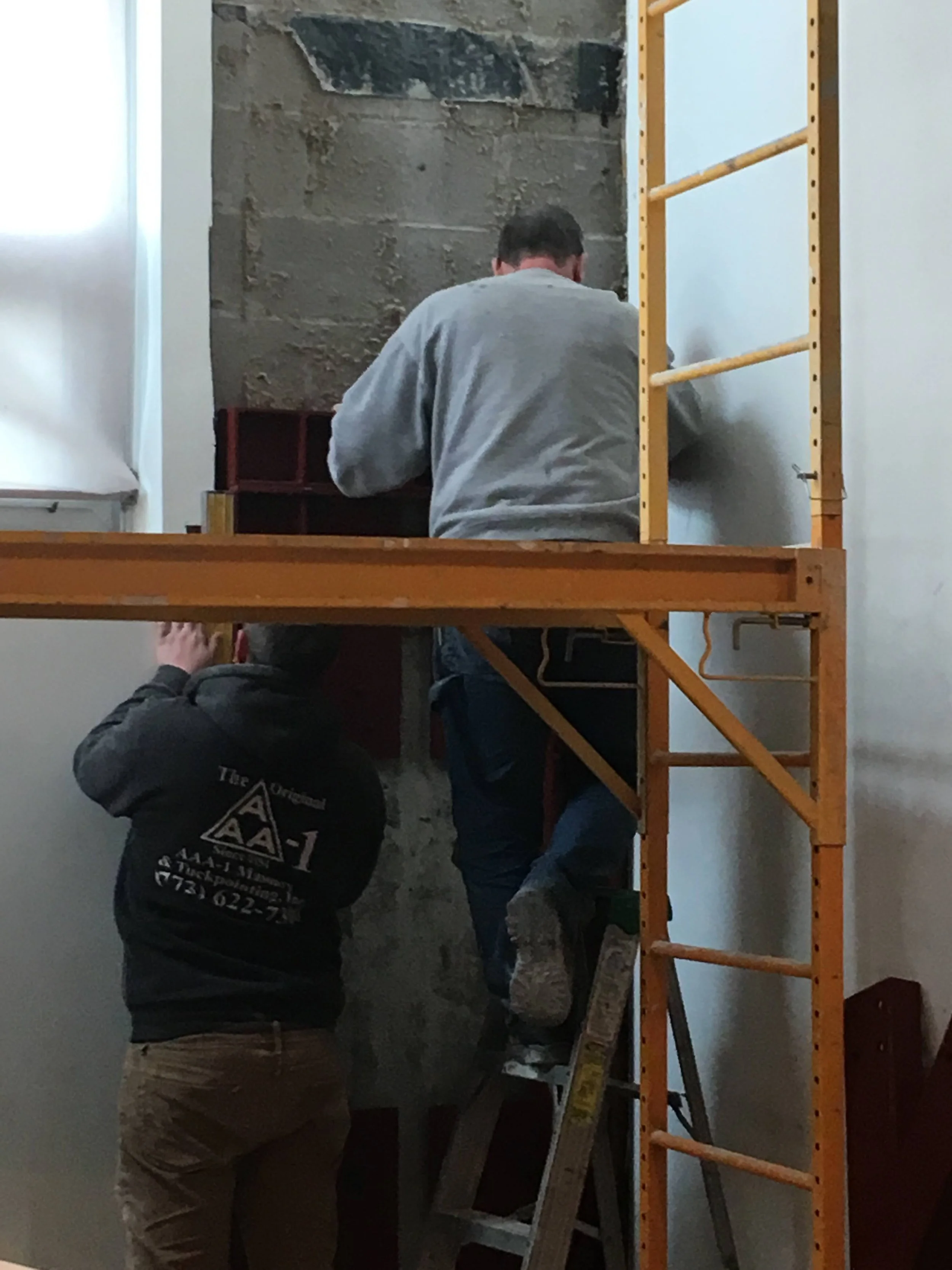 Three men working on a wall in an indoor construction or renovation project. One man is standing on a ladder, another is kneeling, and the third is standing with his back to the camera. The wall has exposed concrete and brickwork.