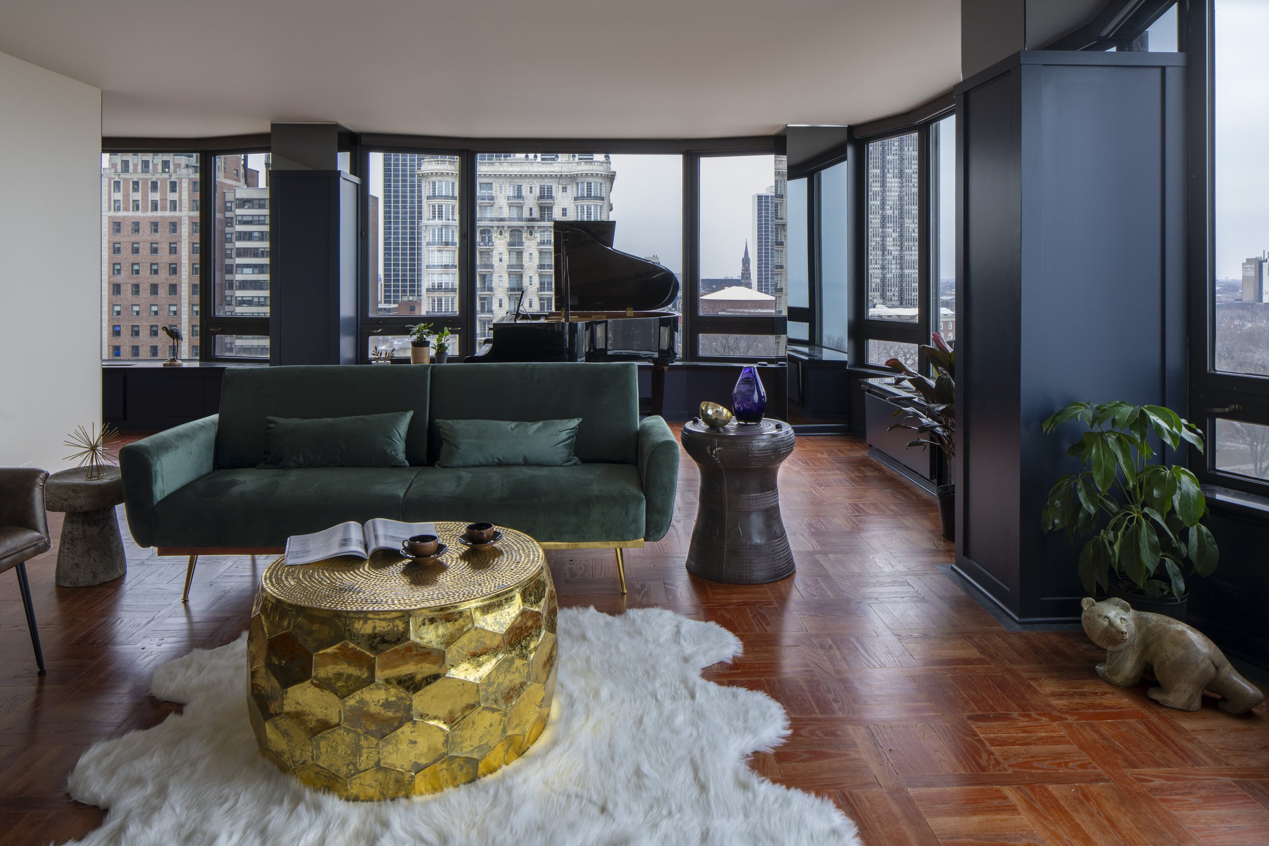 Modern living room with green sofa, white fur rug, gold hexagonal coffee table, and view of city skyline through large windows.