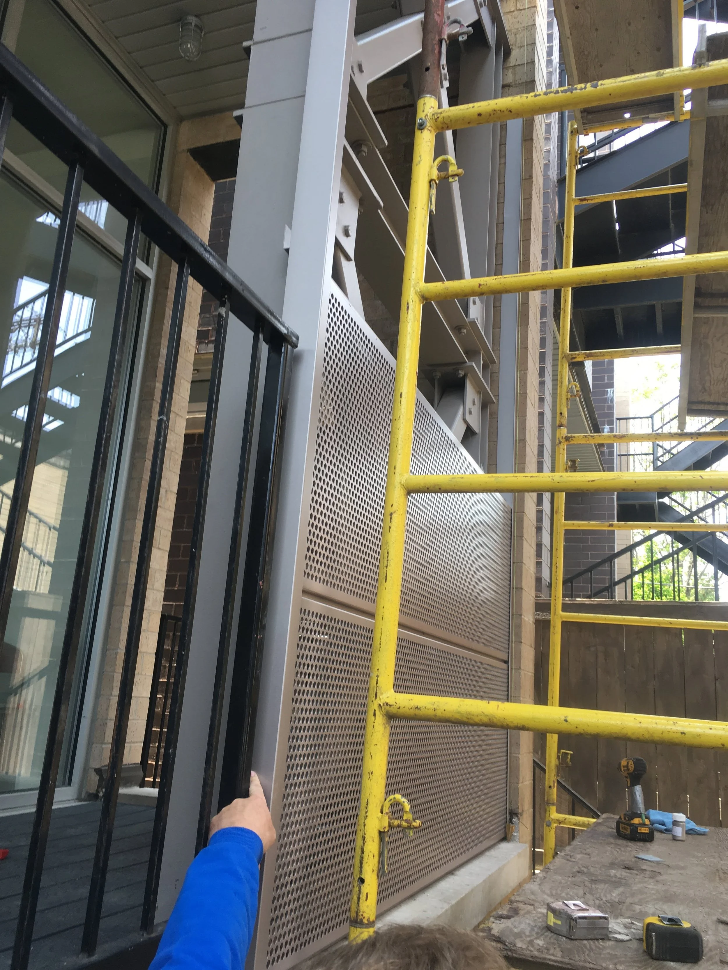 Construction or installation work outdoors showing a person in a blue shirt installing a perforated metal panel on a building exterior, with yellow scaffolding, tools, and power drill nearby.