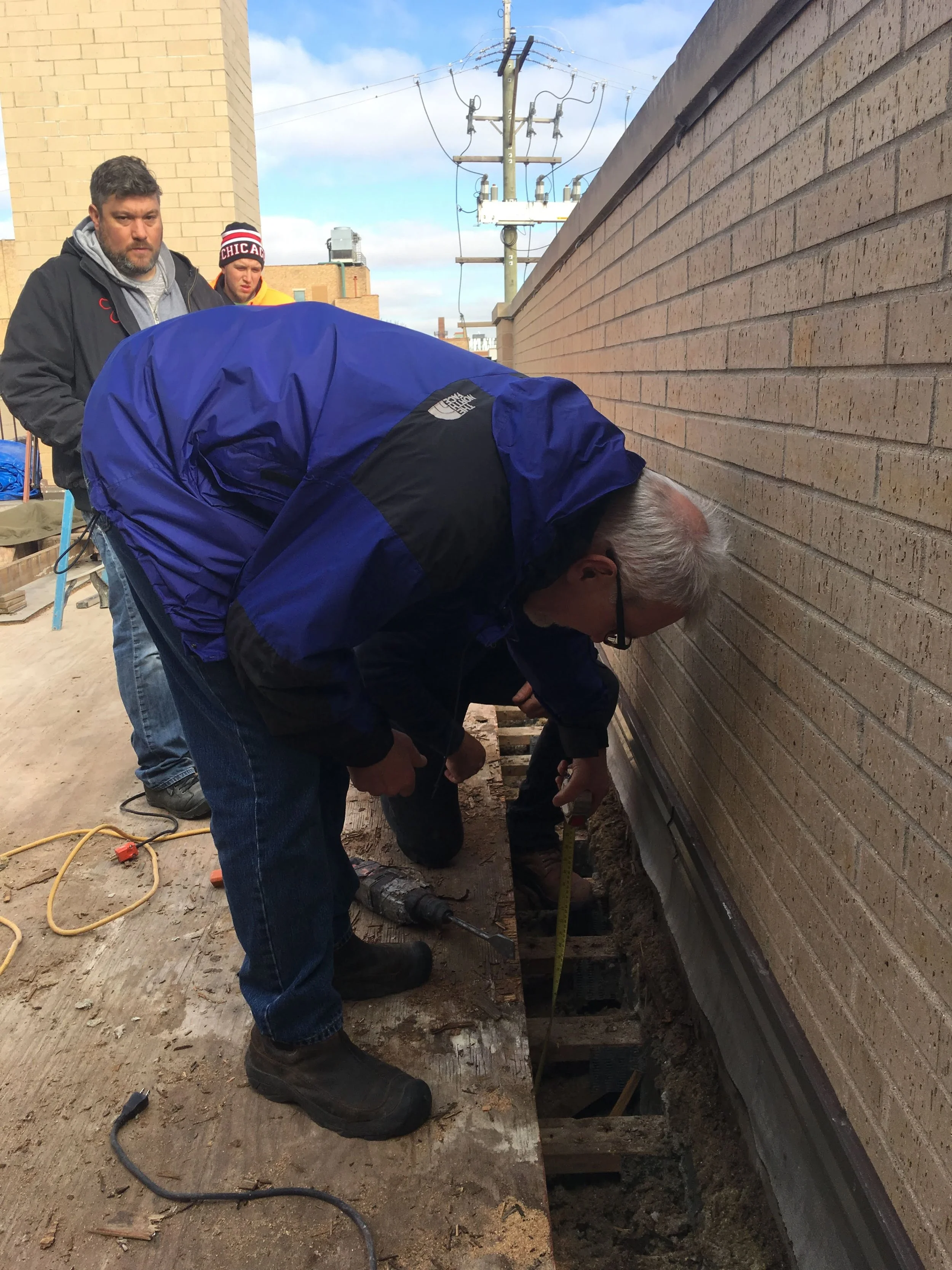 People working on the ground next to a brick building, with one person measuring and others handling tools, during a construction project.