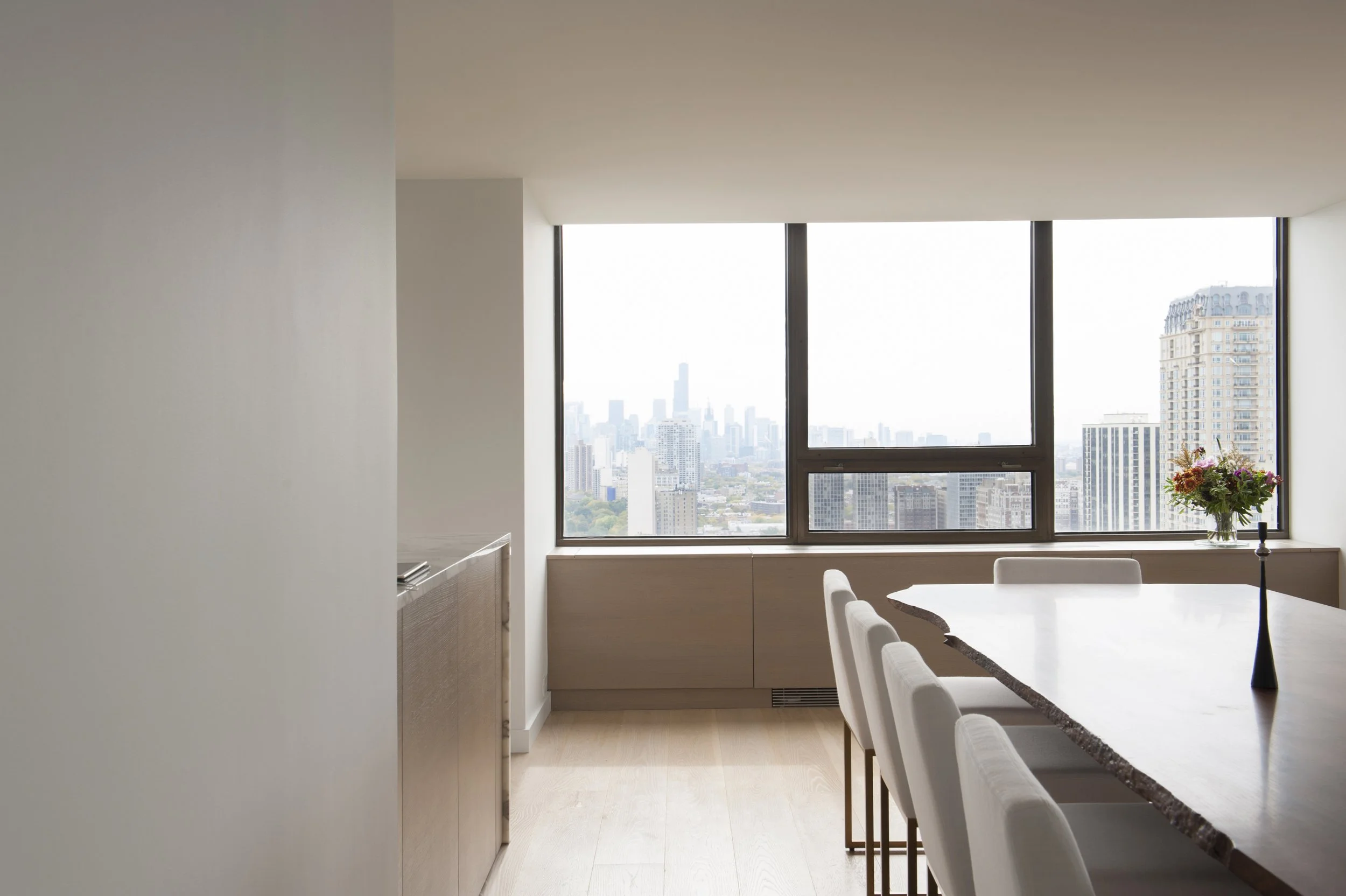 Modern dining room with a large wooden table, white chairs, a bouquet of flowers, and a window view of a city skyline with skyscrapers