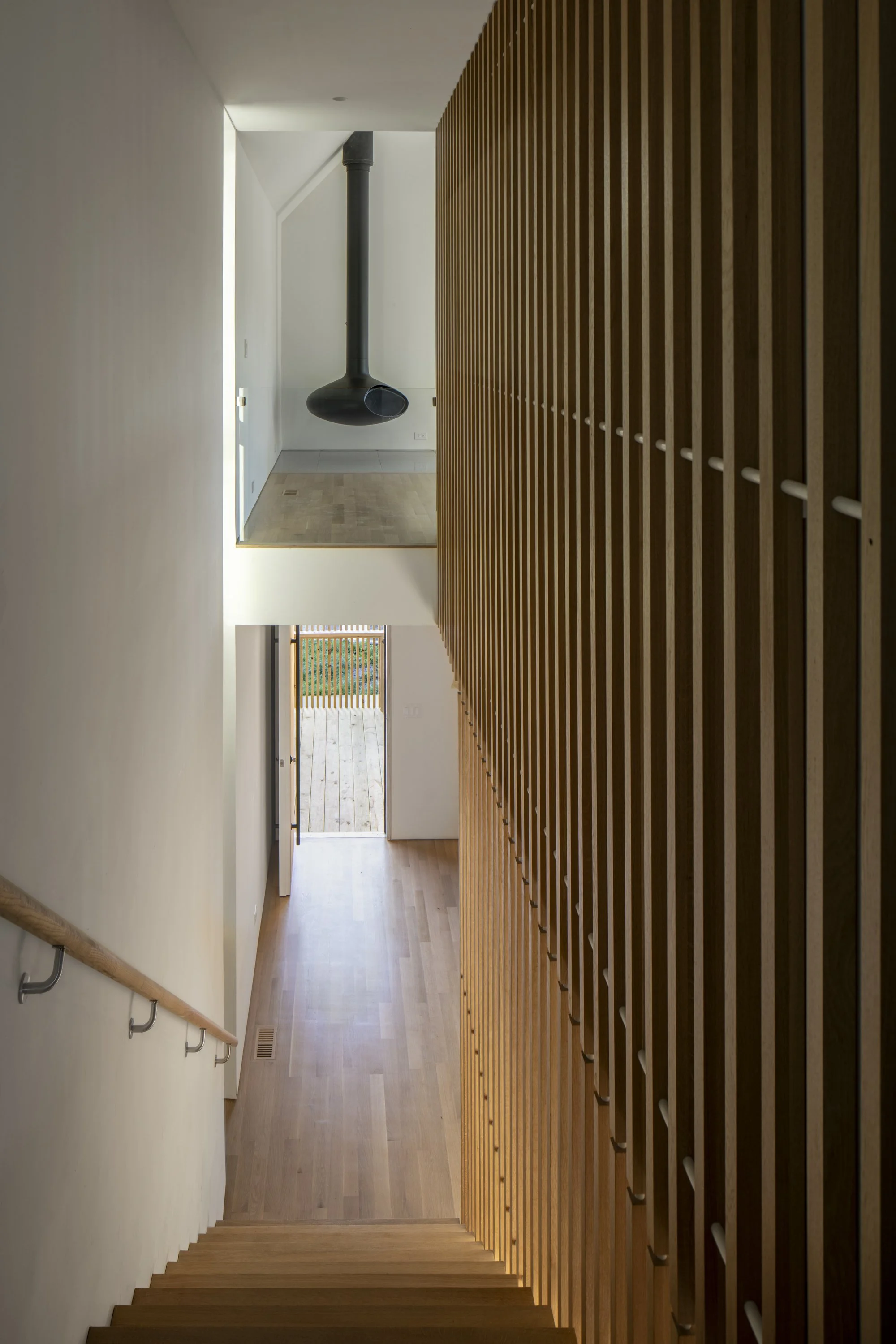 Interior view of a modern house staircase with wood slats on the right side, leading down to a hallway with a door opening to an outdoor deck, featuring minimalistic design and natural light.