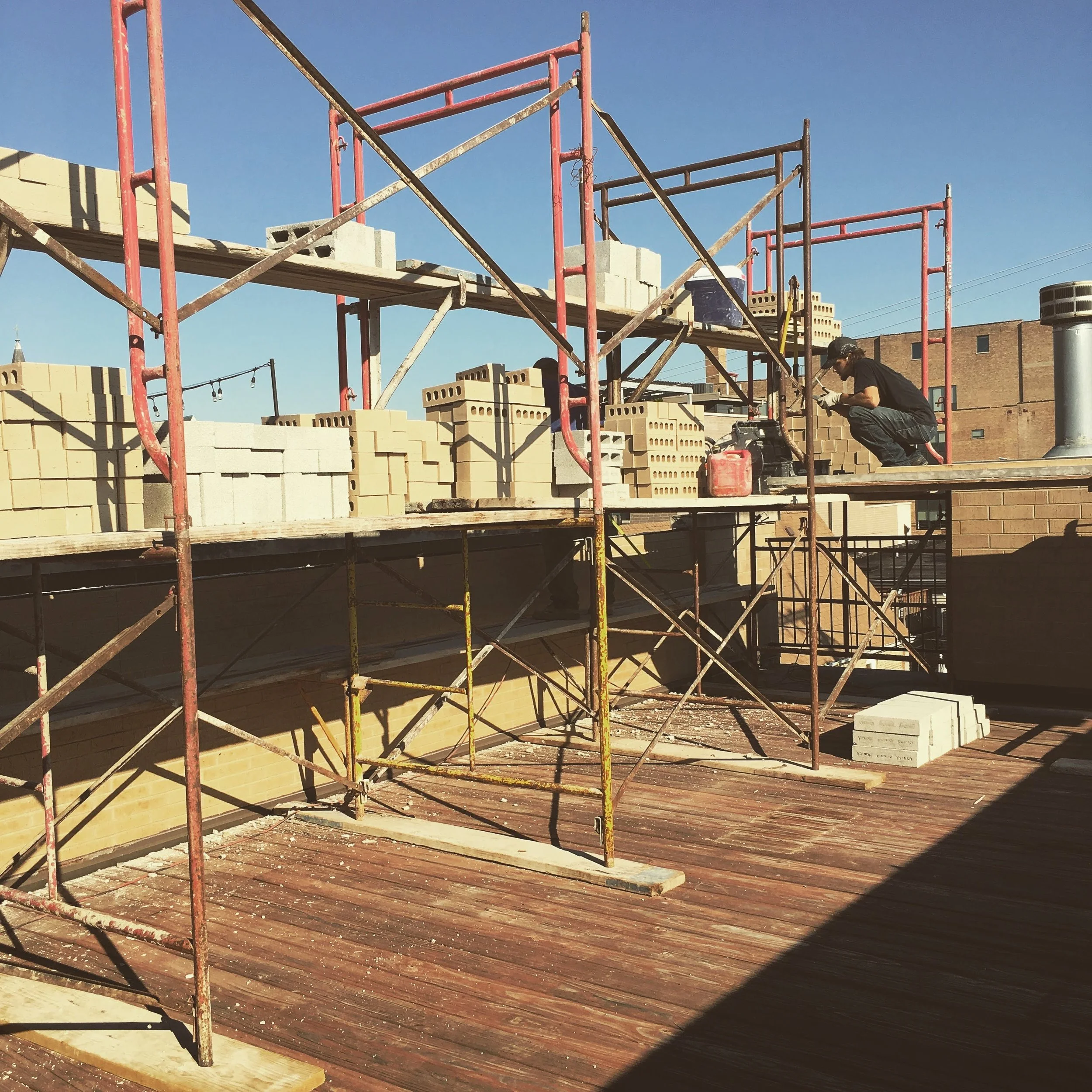 Construction worker on scaffolding working with tools beside a brick building under sunny clear blue sky.