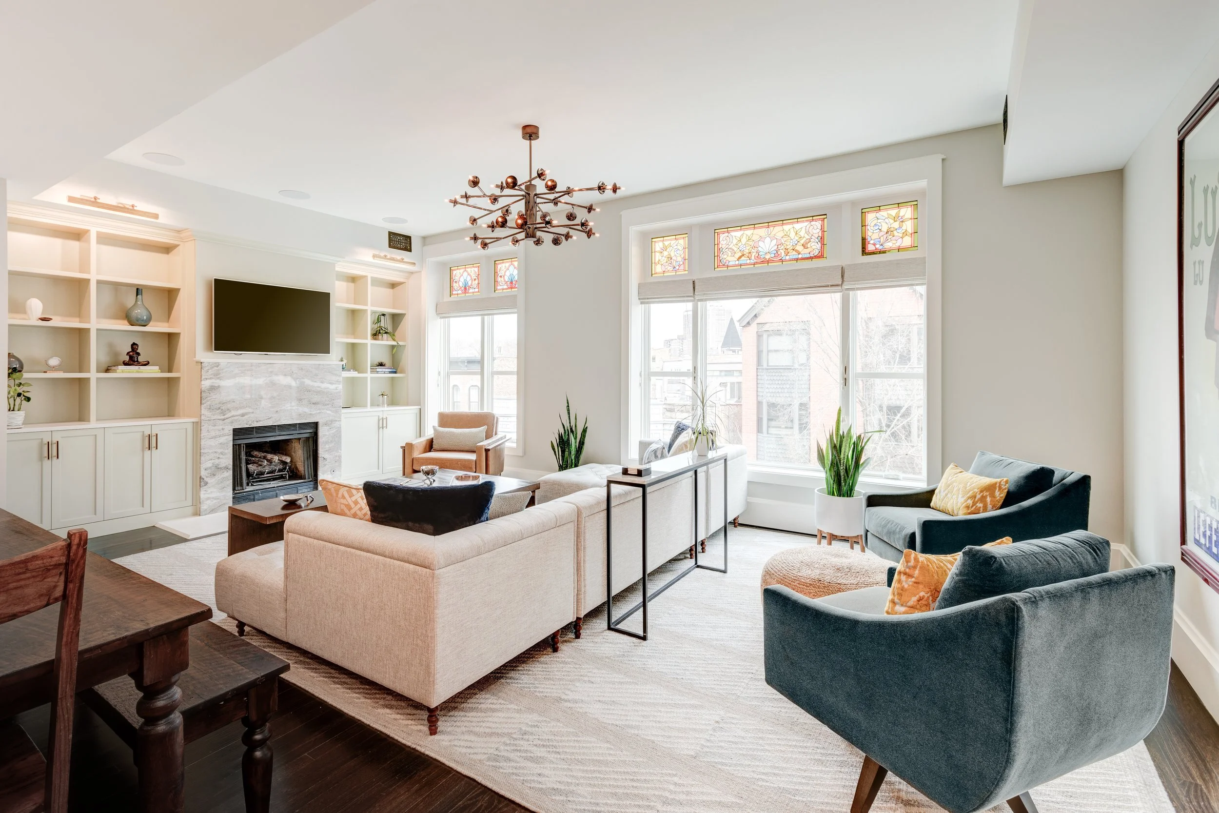 Bright living room with large windows, an off-white sofa, two blue armchairs with pillows, a patterned rug, a modern chandelier, and built-in shelves surrounding a fireplace with a TV above.