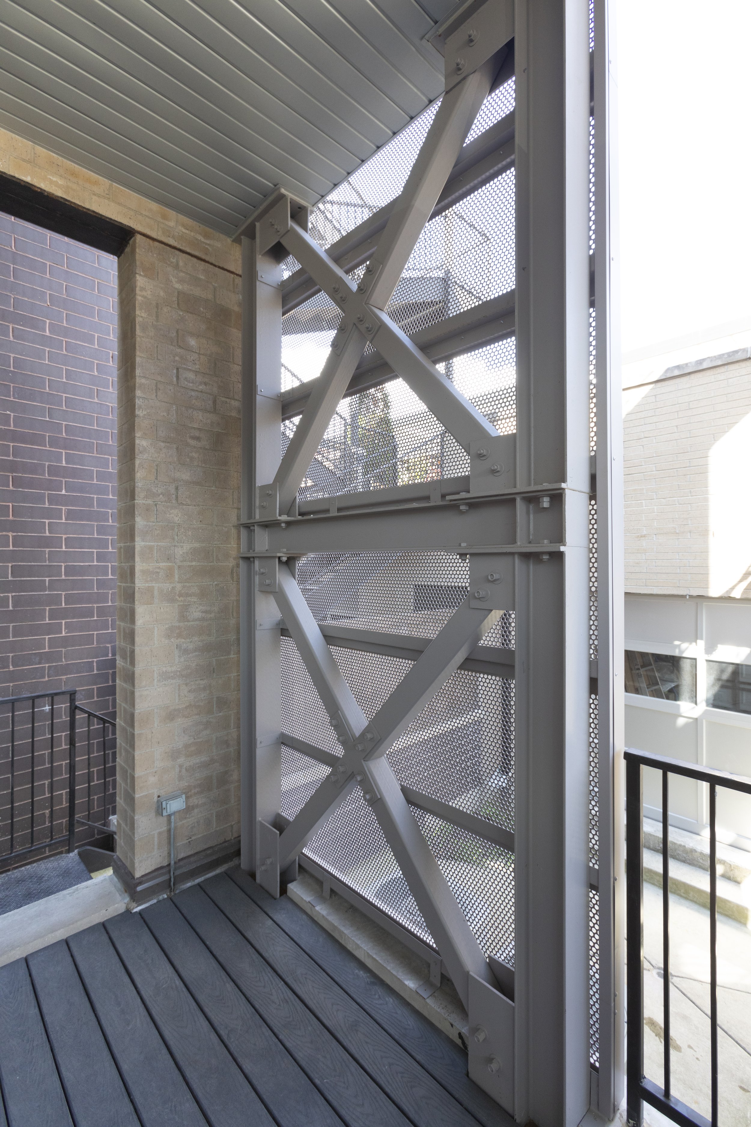 Exterior shot of a metal fire escape ladder with a grated surface, attached to a brick wall on a balcony or fire exit area.