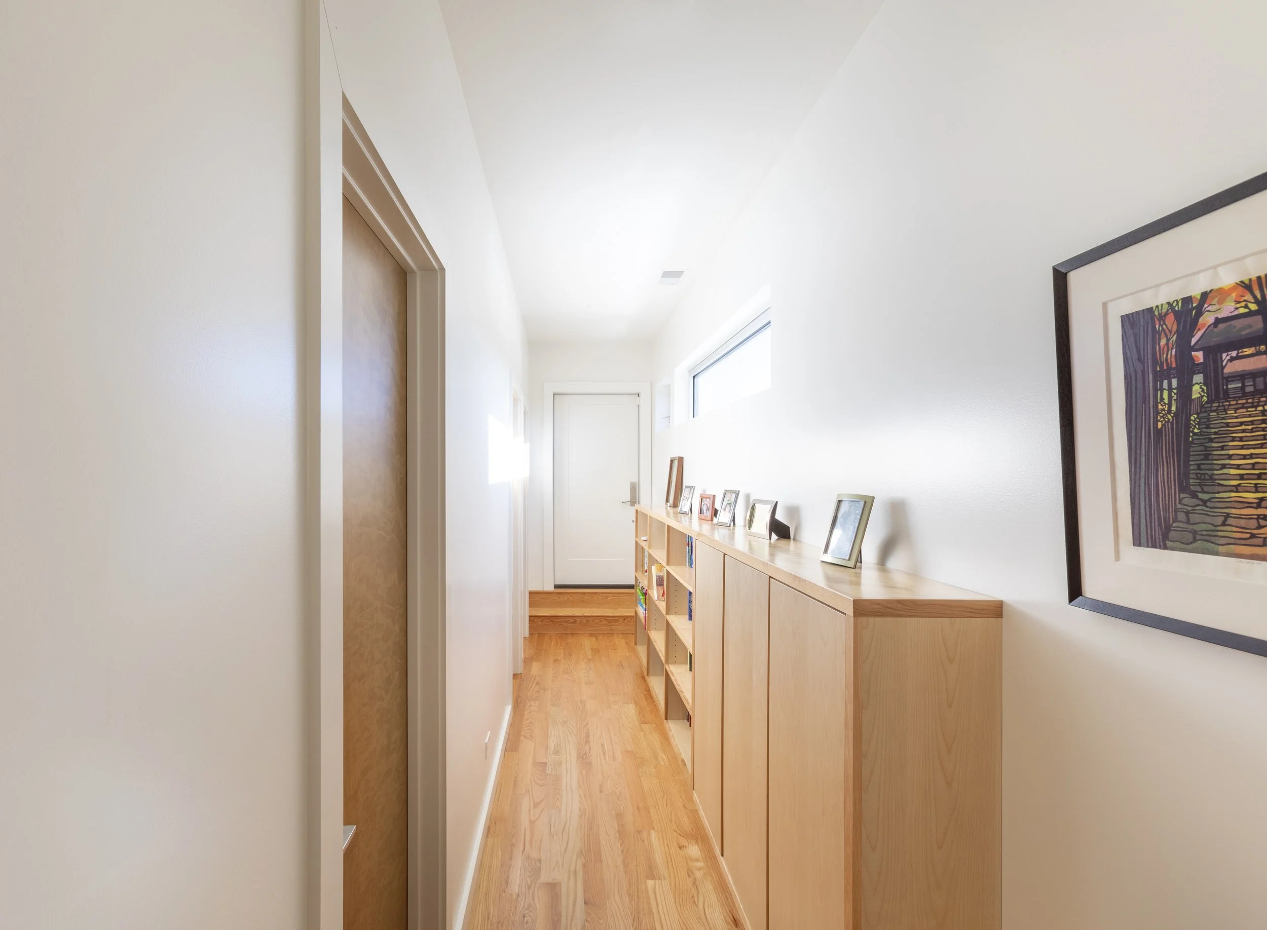 Hallway with wooden floor, white walls, a wooden cabinet with framed photos, and artwork on the wall.