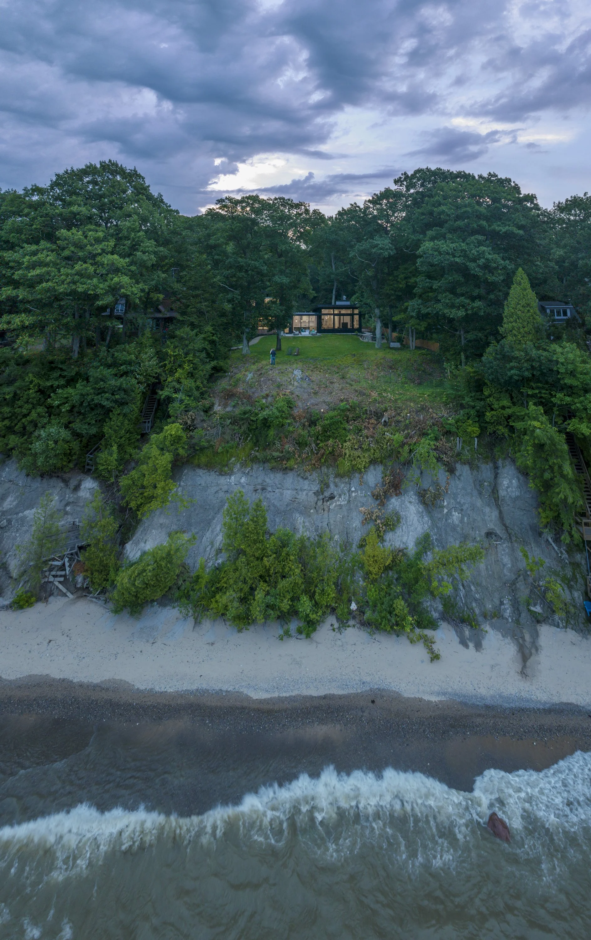 Aerial view of a house built on a cliff overlooking the beach with waves crashing below, surrounded by lush green trees and a cloudy sky.