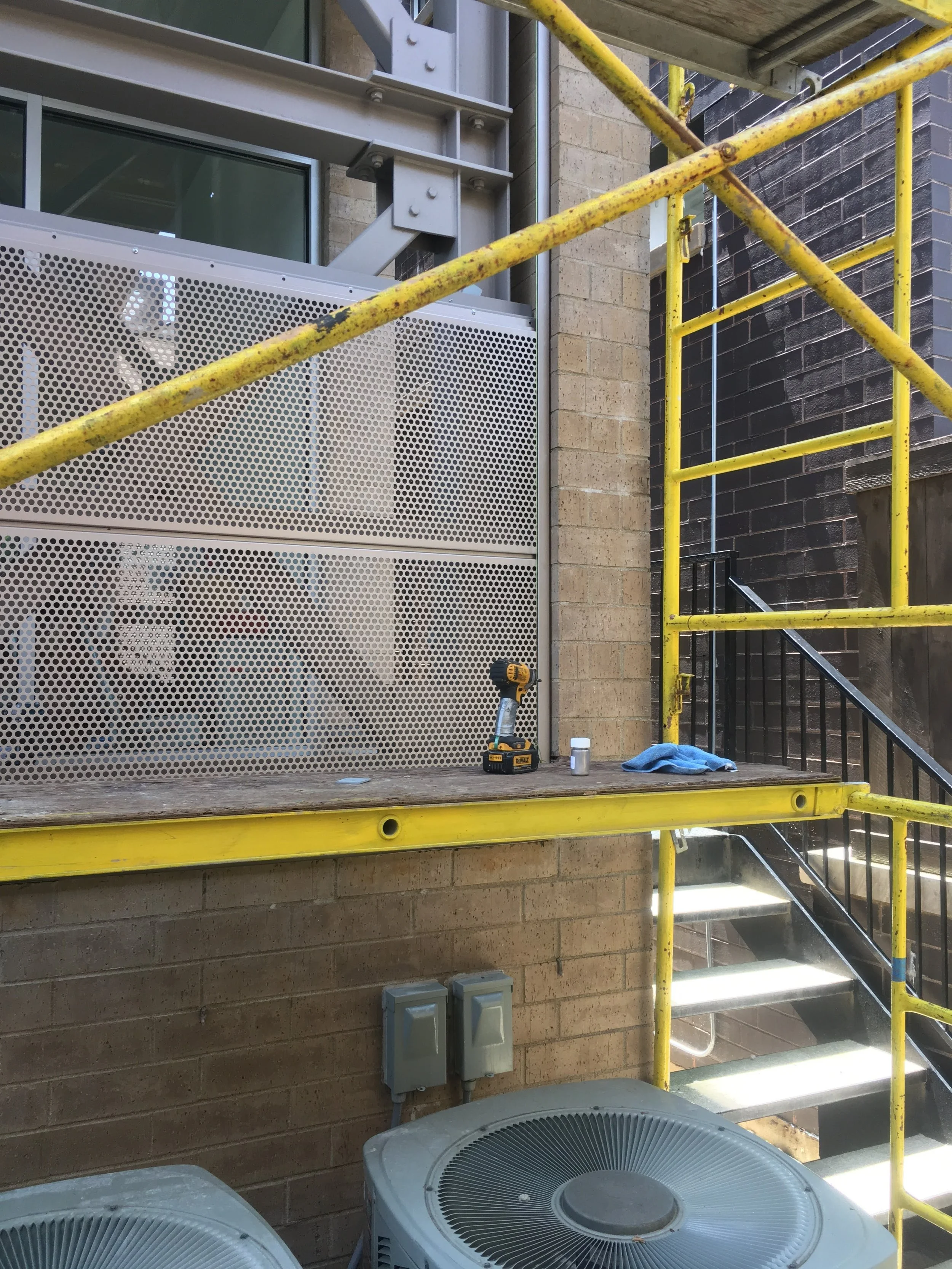 Construction scaffolding with yellow railings outside a brick and metal building, air conditioning units, and some tools.