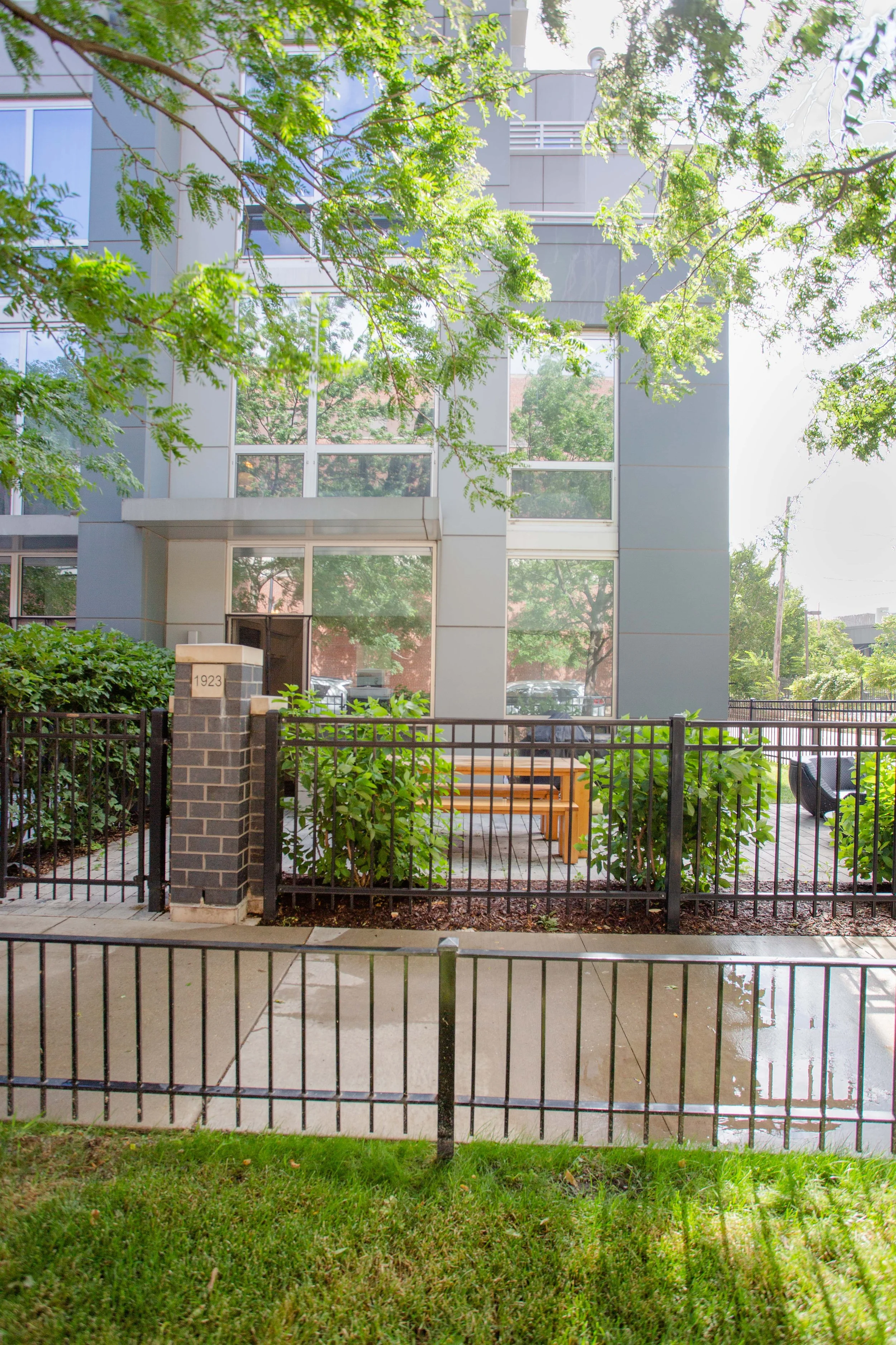 A modern multi-story building with large glass windows, surrounded by green trees and bushes, with a small courtyard that has benches and a black metal fence.