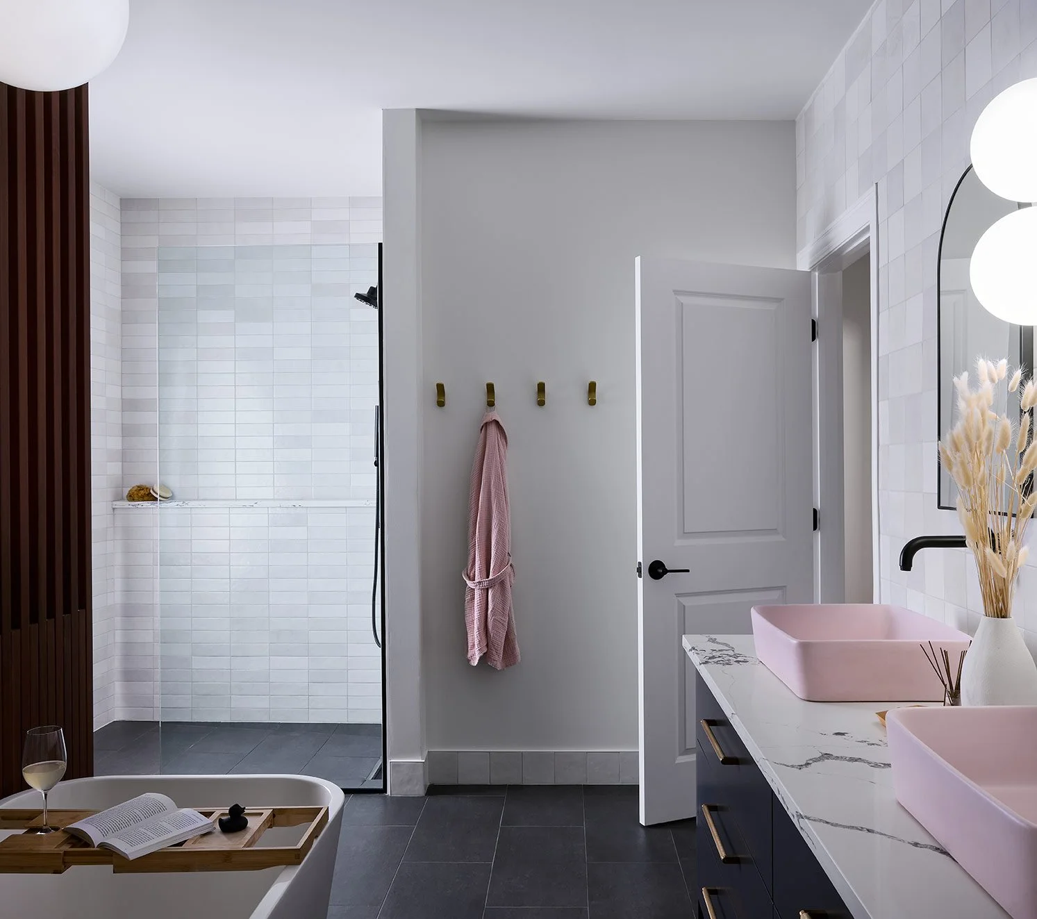 Modern bathroom with a walk-in shower, pink sinks, a marble countertop, and a door leading to the shower area. There is a wooden tray with a glass of wine and an open book on a bathtub, and a pink towel hanging on hooks on the wall.