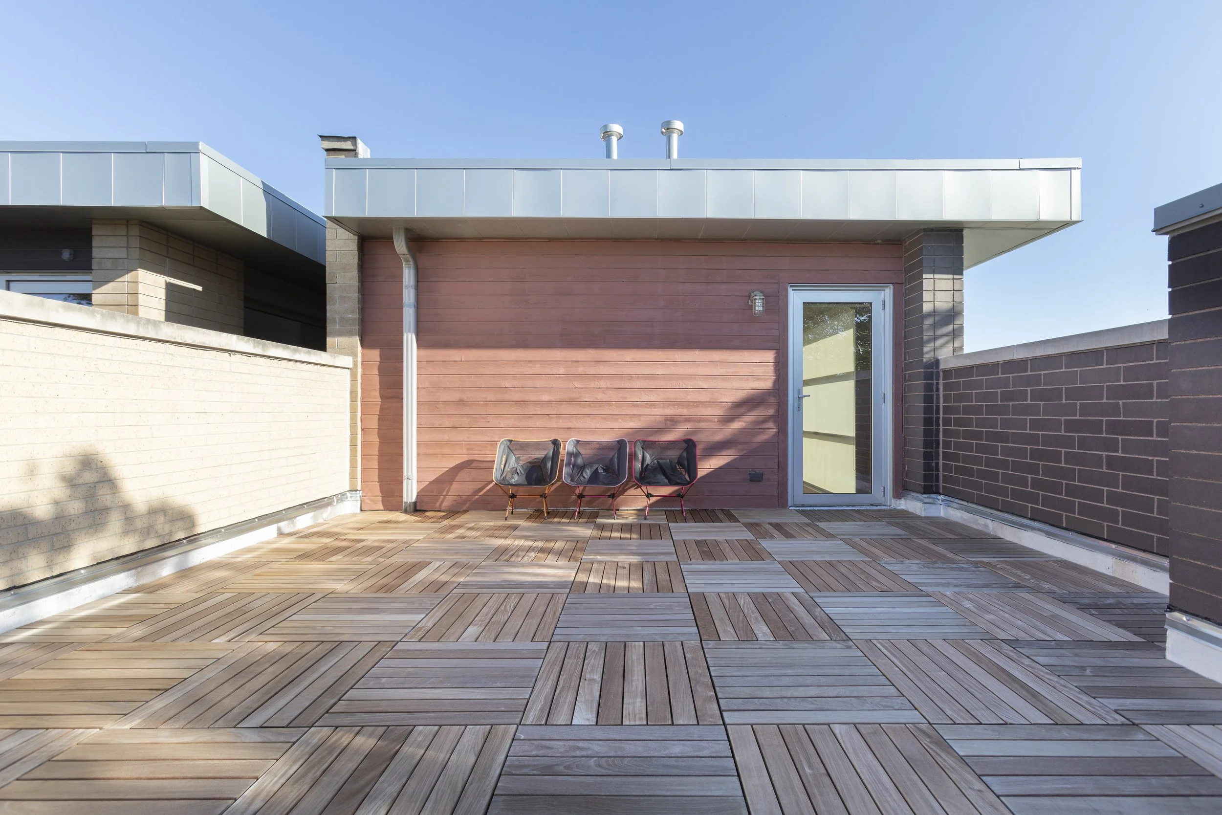 A rooftop balcony with wooden flooring, three chairs, a pink section of wall, a glass door, and brick walls on either side under a clear blue sky.