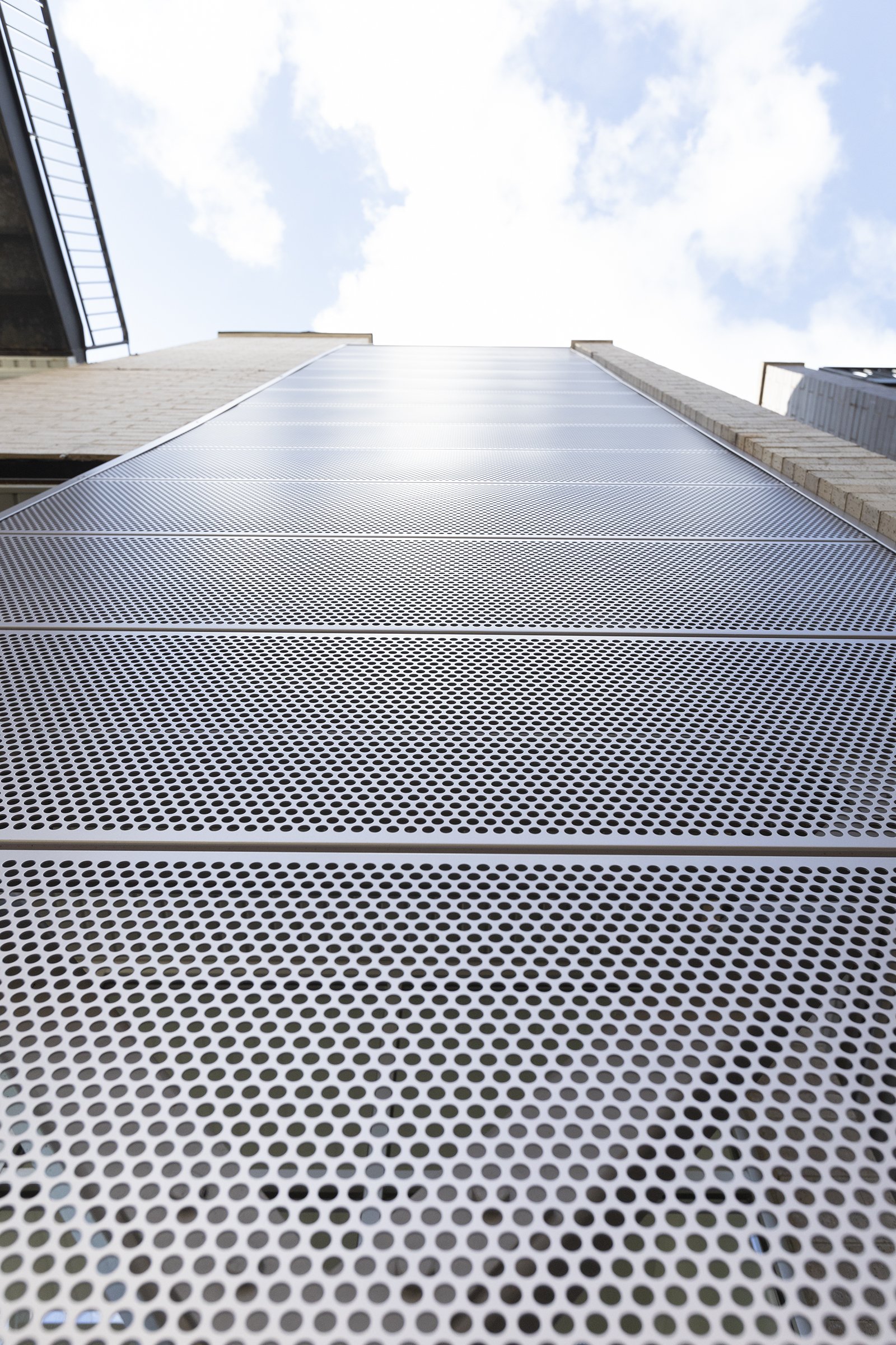 Looking up at a modern building with a perforated metal facade, with a partly cloudy sky above.