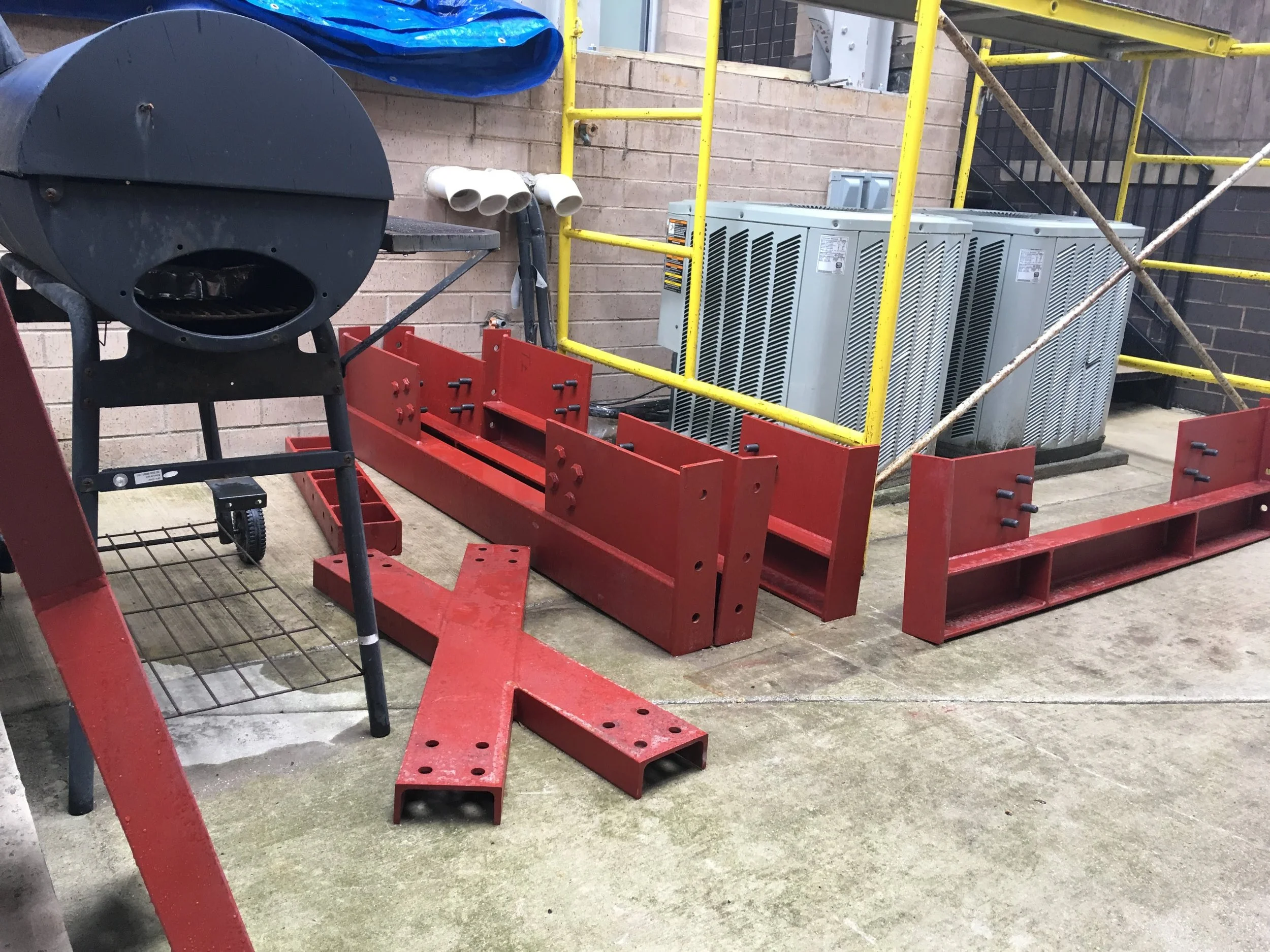 Construction site with red and yellow metal framing, outdoor HVAC units, a barbecue grill, and scaffolding.