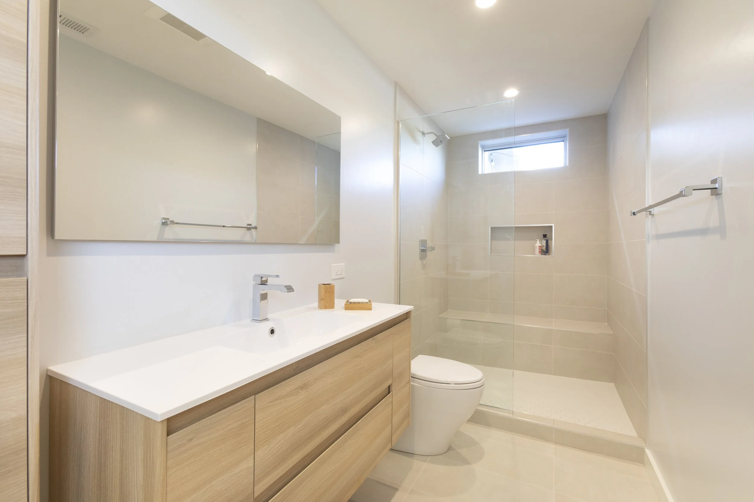 A modern bathroom with a large mirror, a white sink with wooden cabinetry, a toilet, and a walk-in shower with a glass partition. The shower has a small niche with toiletries and a small window near the ceiling.