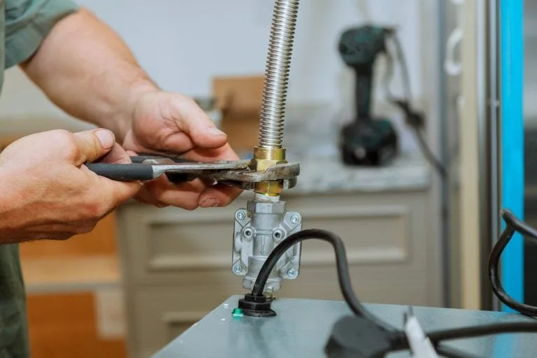 Person using a wrench to work on a mechanical assembly with a flexible metal spring, with a cordless drill and workspace in the background.