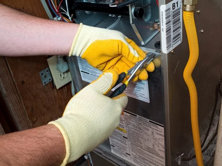 Person wearing yellow work gloves using a screwdriver on a metallic air handler or furnace unit.