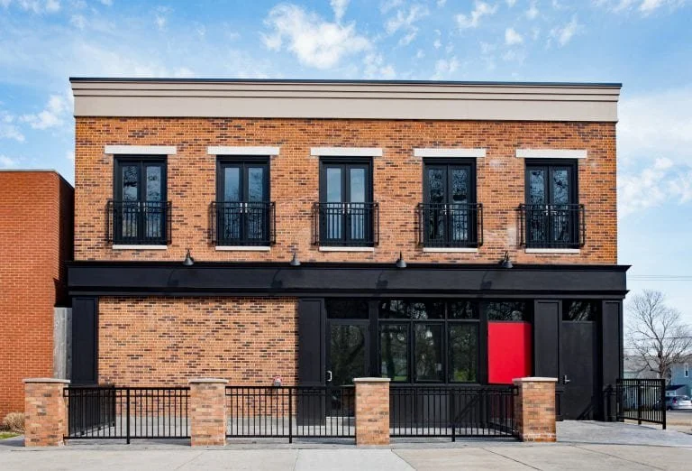 Two-story brick building with black metal balcony railings on the upper floor windows and a black facade on the ground floor, including a red door.