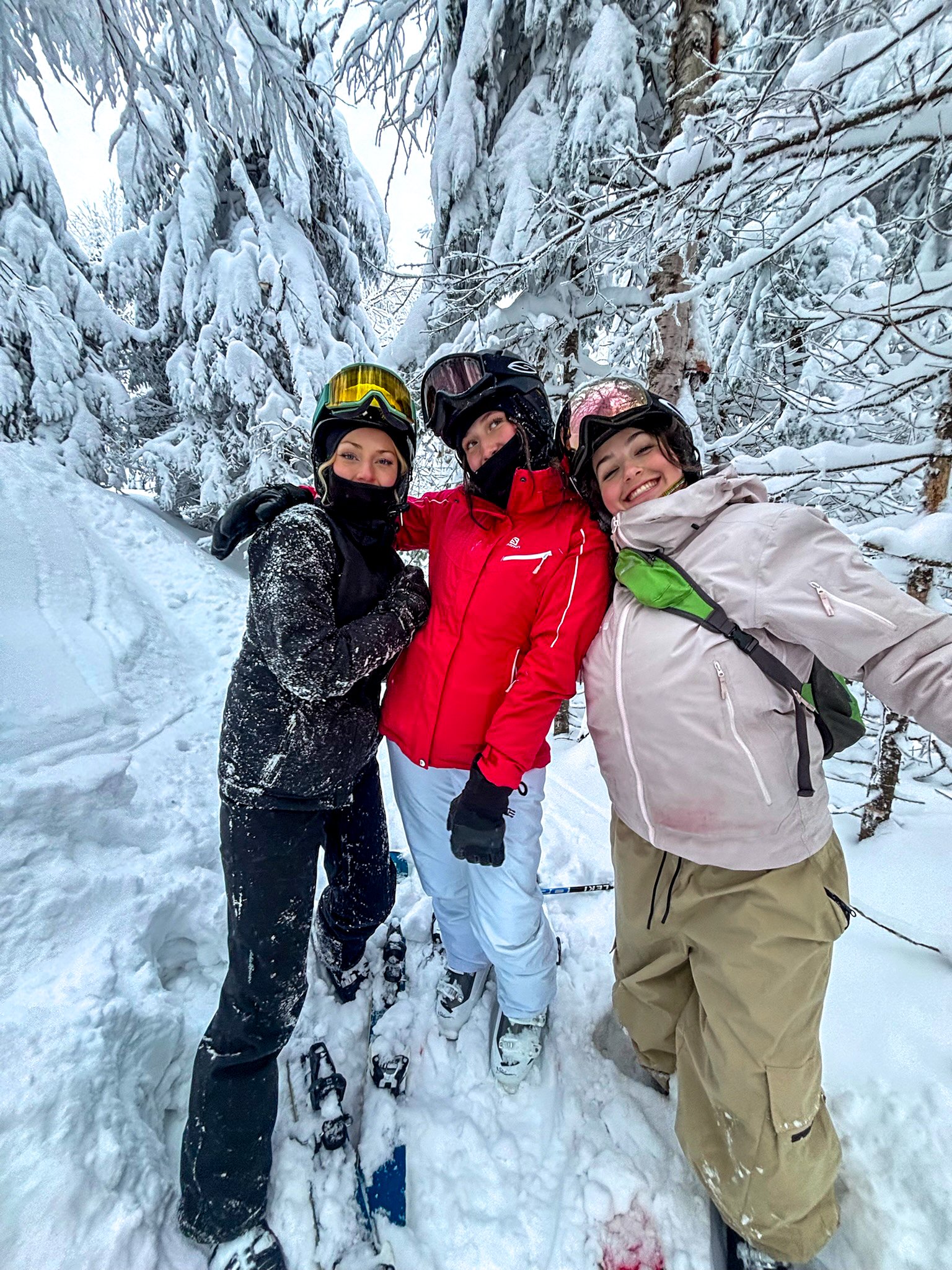 Three women wearing winter sports gear, including helmets, goggles, and snow jackets, posing together in a snowy forest with snow-covered trees in the background.