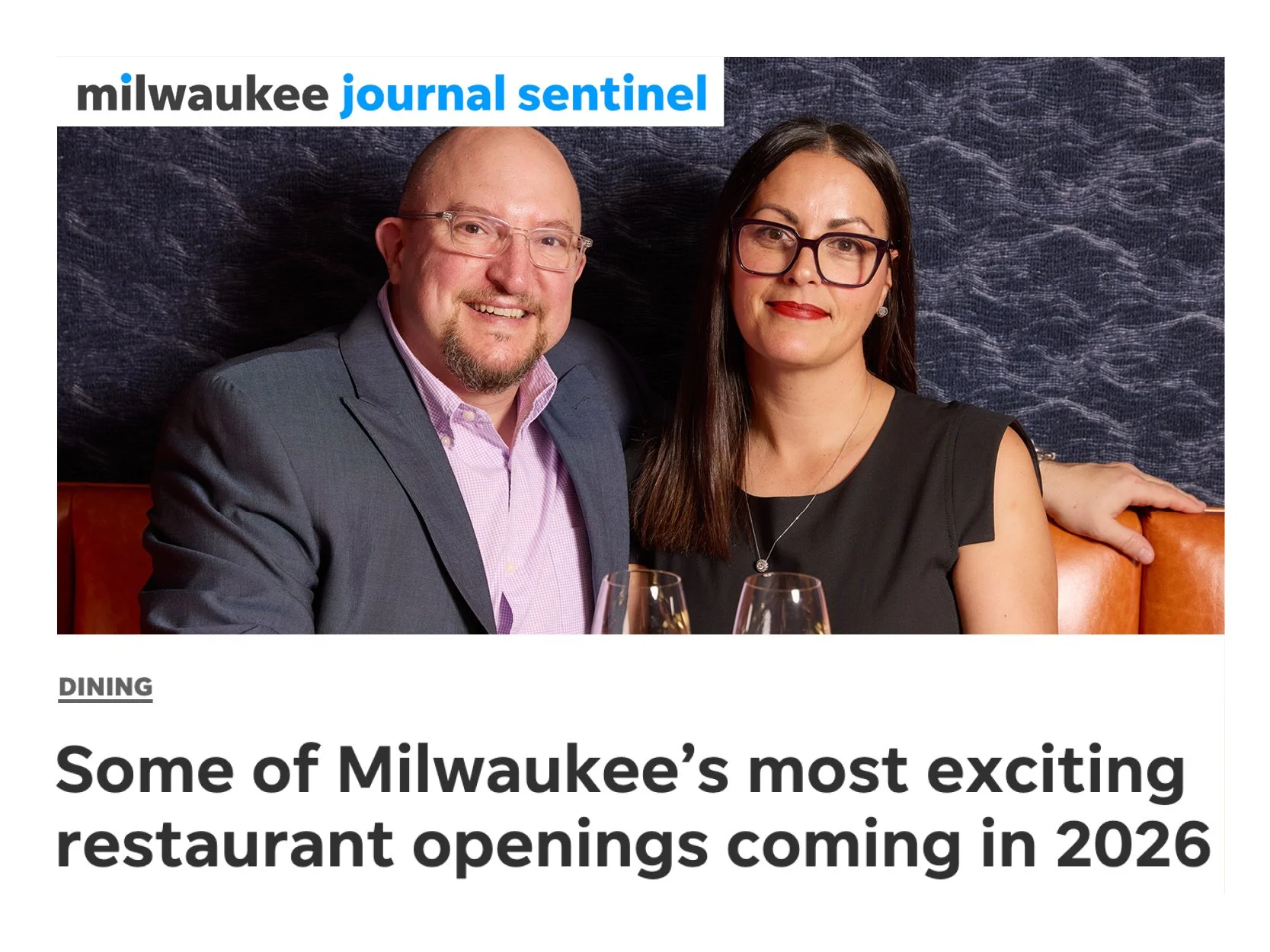 A man and woman sitting together at a restaurant, smiling at the camera, with two glasses of wine in front of them.