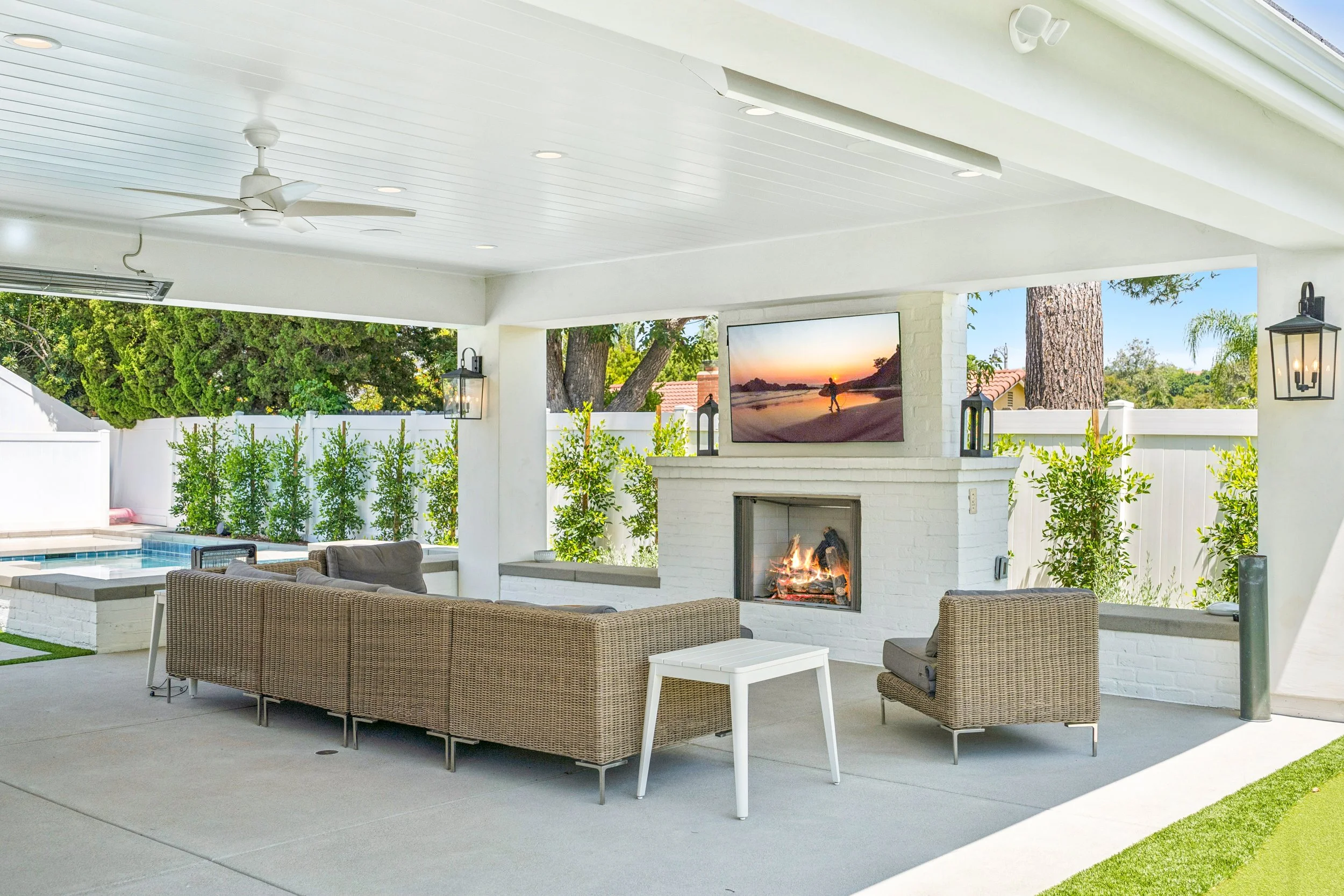 Covered outdoor living area with white brick fireplace, mounted TV, lounge seating, pool views, ceiling fan, and indoor-outdoor access from the home.