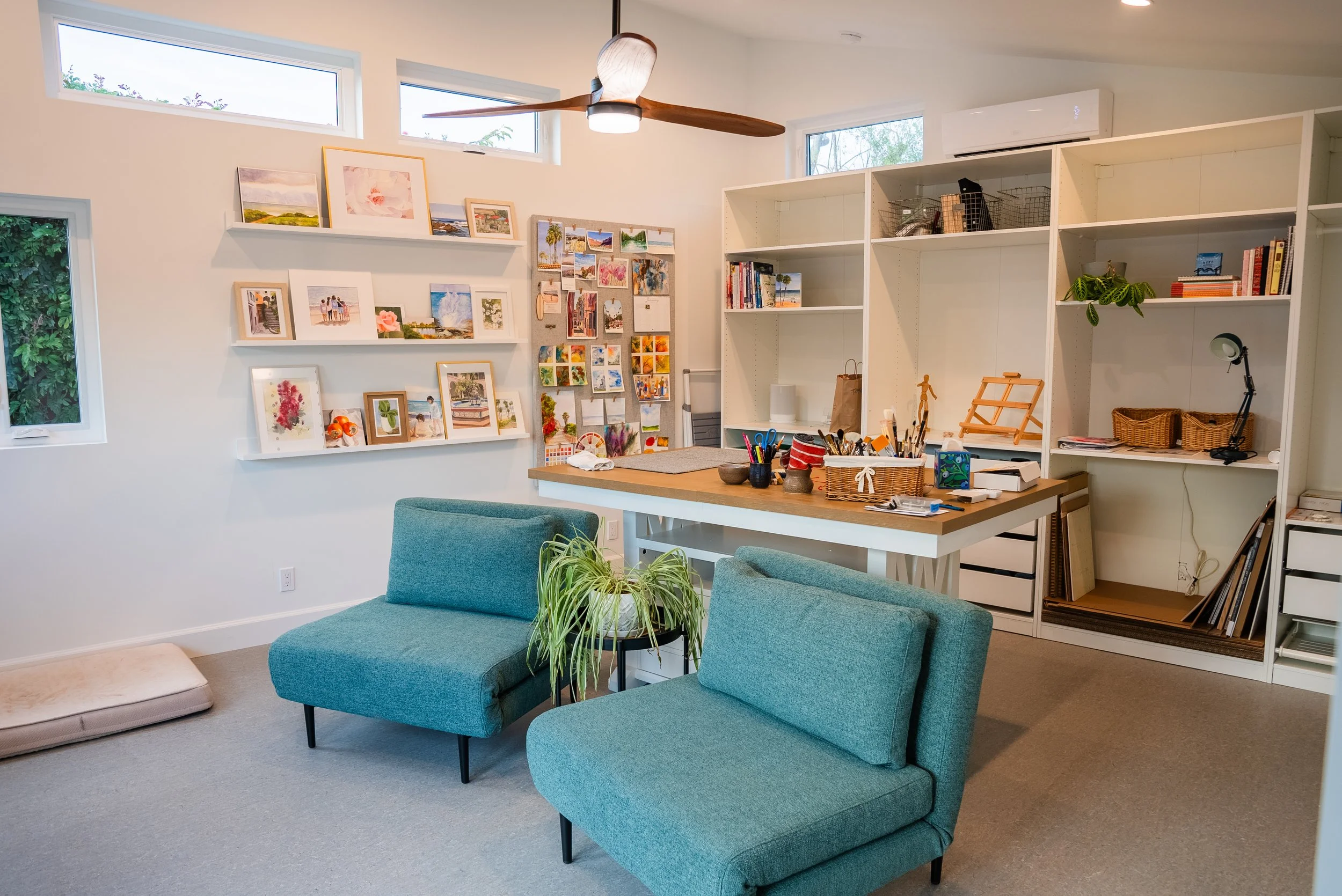 Light-filled ADU art studio with teal lounge chairs, high windows, floating shelves, storage cabinets, worktable, and ceiling fan.