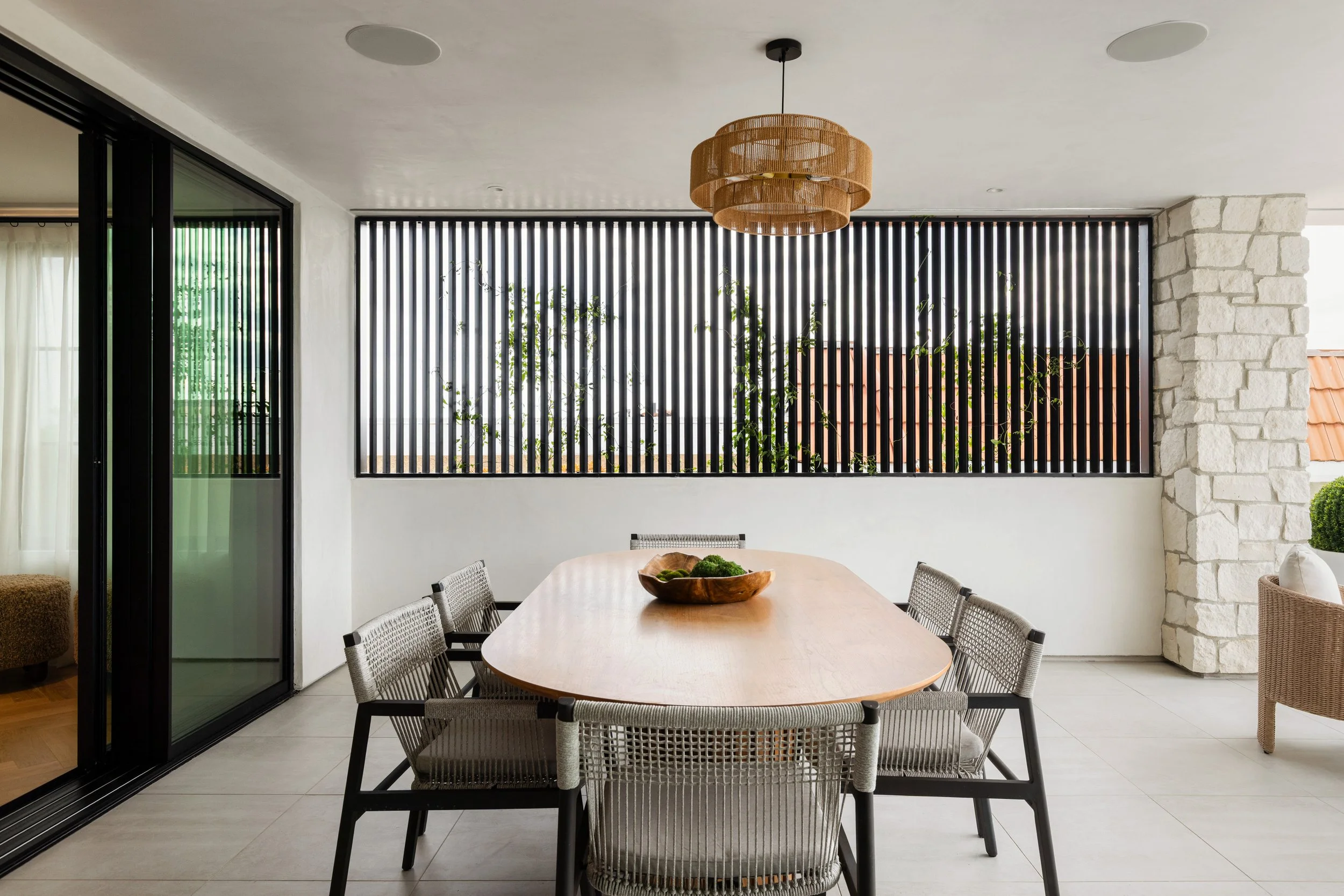 Rear exterior of a modern coastal home with glass balcony, stone columns, black framed doors, outdoor seating, and warm evening lighting.