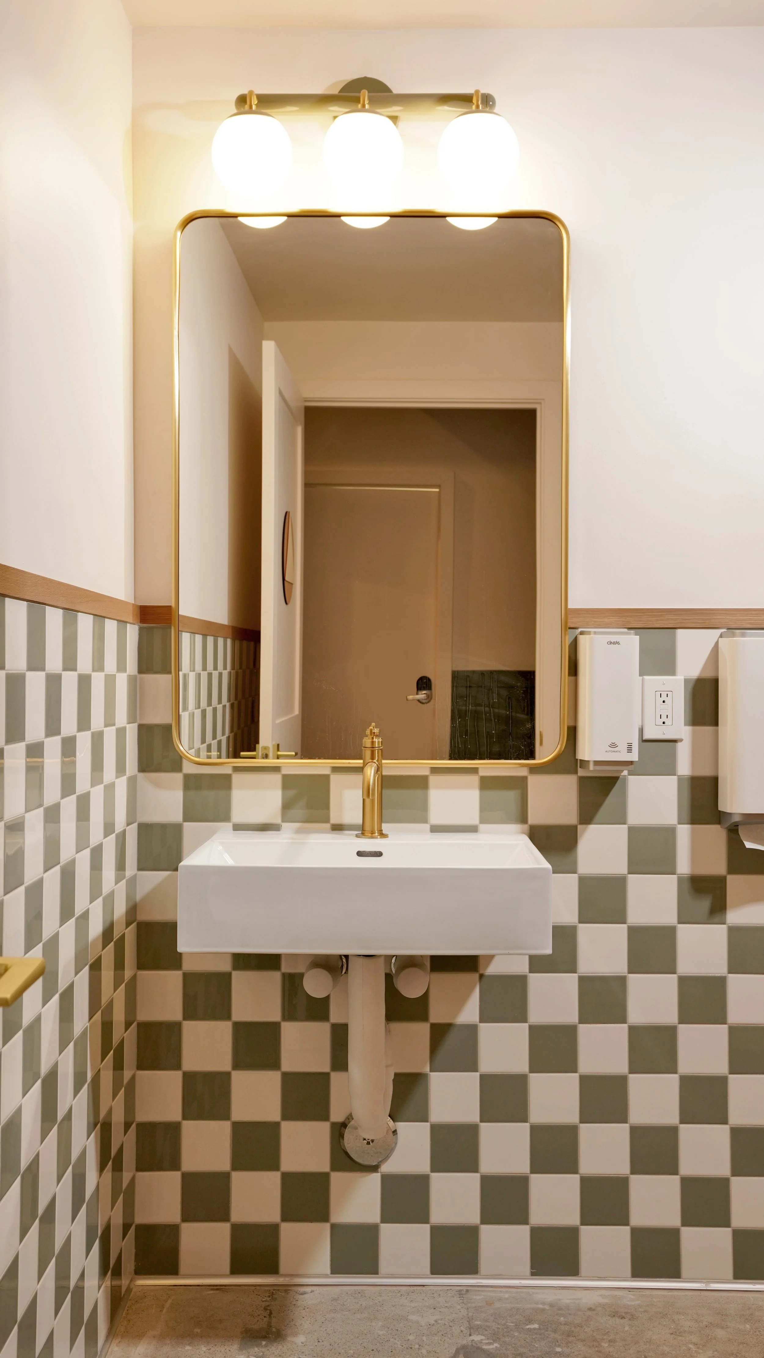 Bathroom vanity with checkerboard tile, wall-mounted sink, brass fixtures, and arched mirror in a completed commercial coffee shop build-out.