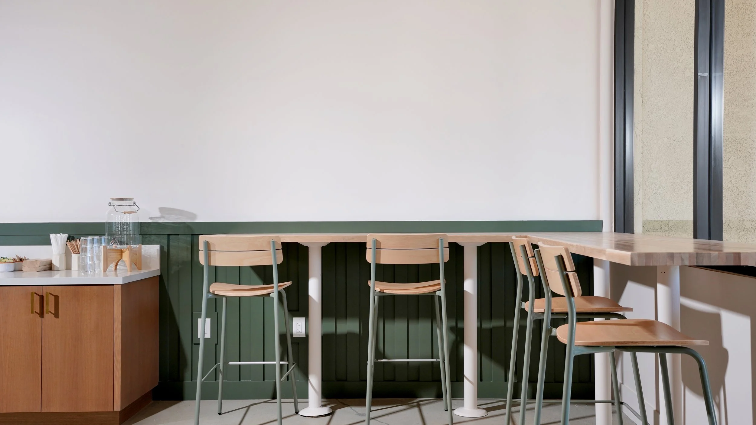 Coffee shop bar seating with light wood counter, green metal stools, wood cabinetry, and black window framing.